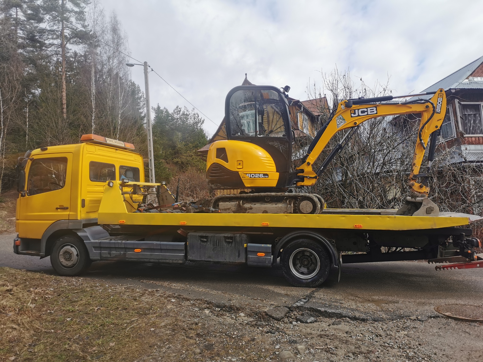 Żółty transportowiec z mini koparką JCB 8026 CTS na platformie, na tle drzew i domu. Usługa transportu maszyn budowlanych.