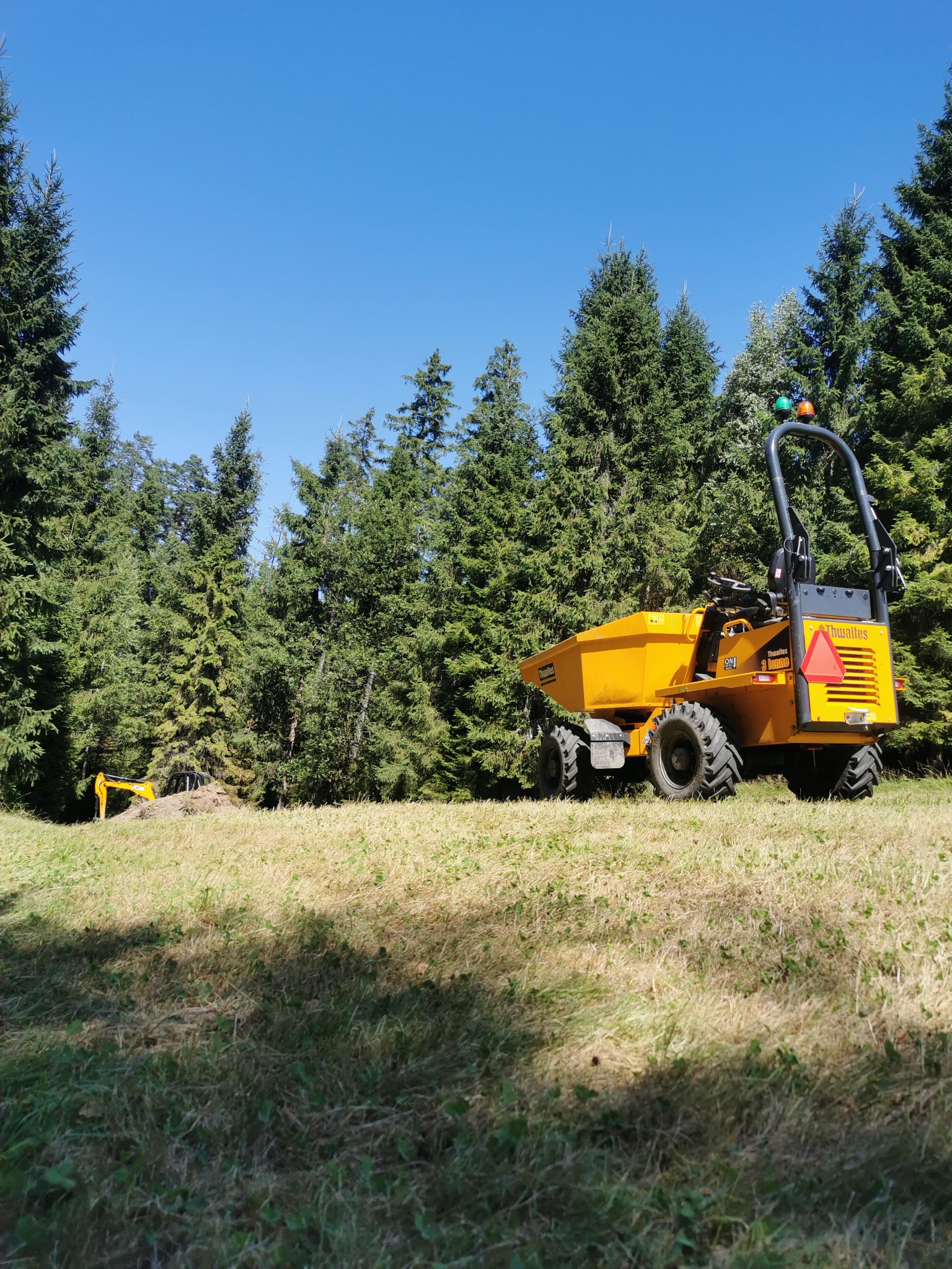 Żółta wywrotka Thwaites 3 tonne na tle lasu iglastego i błękitnego nieba. W tle widoczna koparka. Prace ziemne w toku, słoneczny dzień.