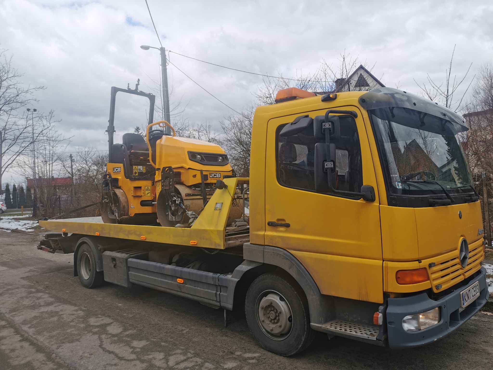 Żółta laweta z walcem drogowym JCB na platformie, widok z boku. Pojazd na szarej, popękanej drodze, w tle drzewa i budynki pod pochmurnym niebem.