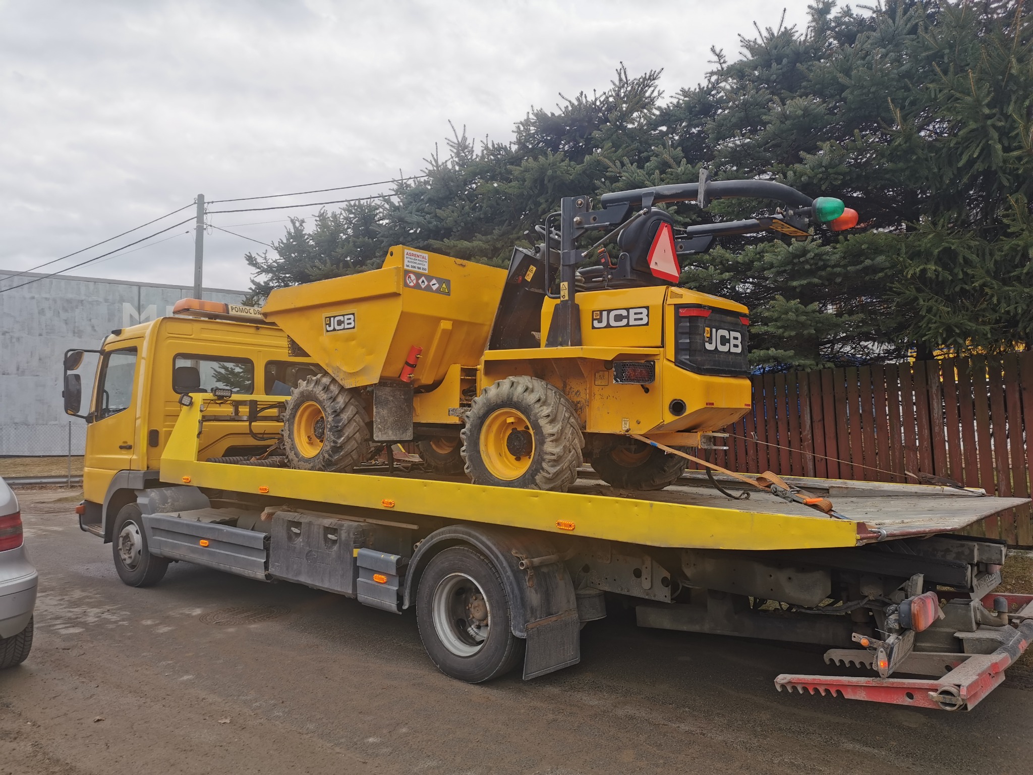 Żółta wywrotka JCB na lawecie, zabezpieczona pasami transportowymi, na tle drzew iglastych i budynku. Transport maszyny budowlanej.