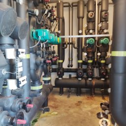 Przedsiębiorstwo Budowlano-Instalacyjne ecoEner Wojciech Jendza - Złożona instalacja grzewcza z rurami, zaworami, manometrami i zielonymi pompami obiegowymi. Widoczne narzędzia i elementy montażowe na posadzce.