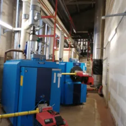 Dwa niebieskie, przemysłowe kotły Buderus z czerwonymi palnikami w kotłowni. Widoczne rury, instalacje i oświetlenie. Ściany częściowo wykończone, sufit z drewnianą konstrukcją.