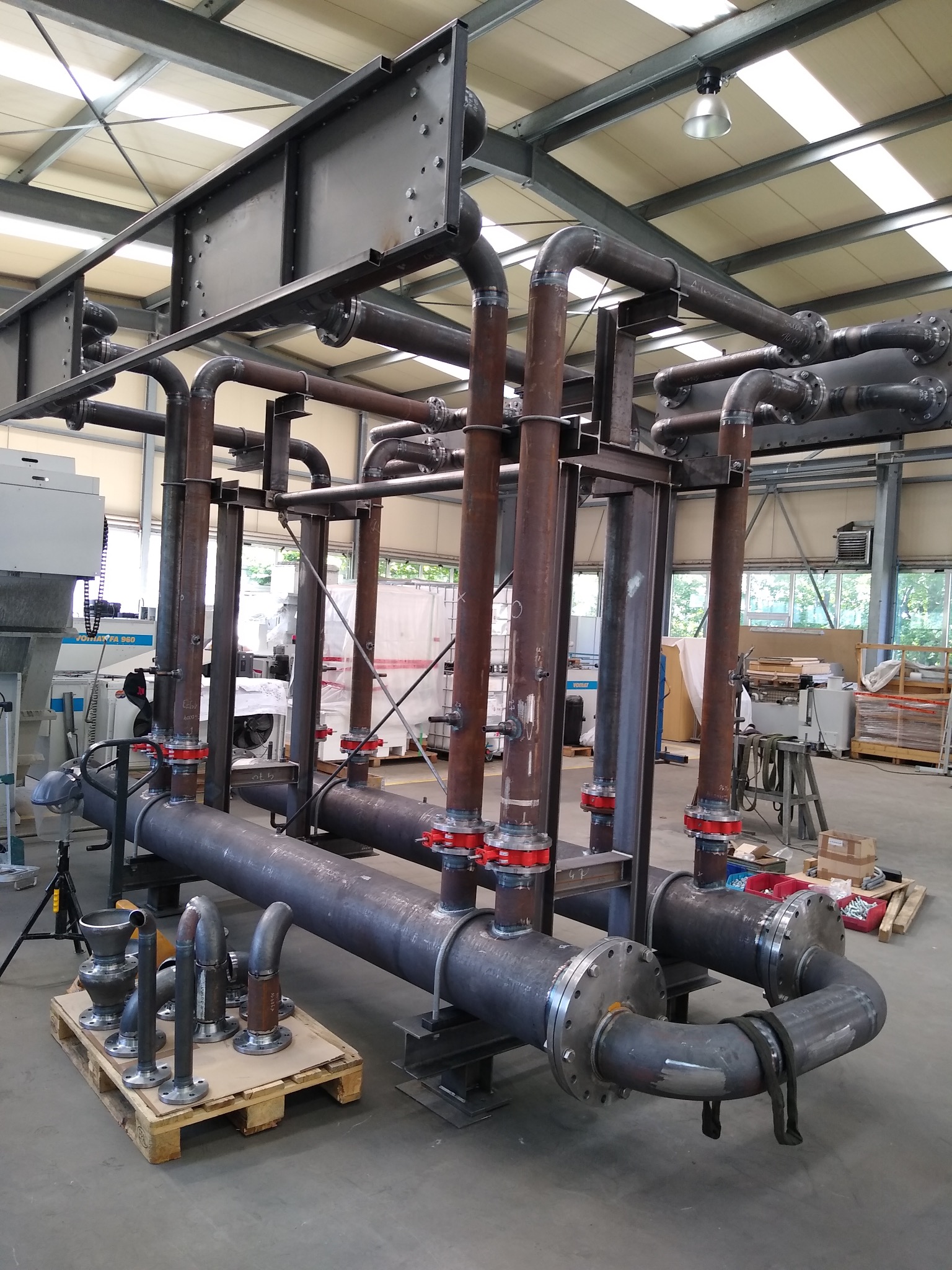 Metalowa konstrukcja instalacji przemysłowej z rurami, złączkami i spawami, widoczna w hali produkcyjnej. Elementy na palecie gotowe do montażu.