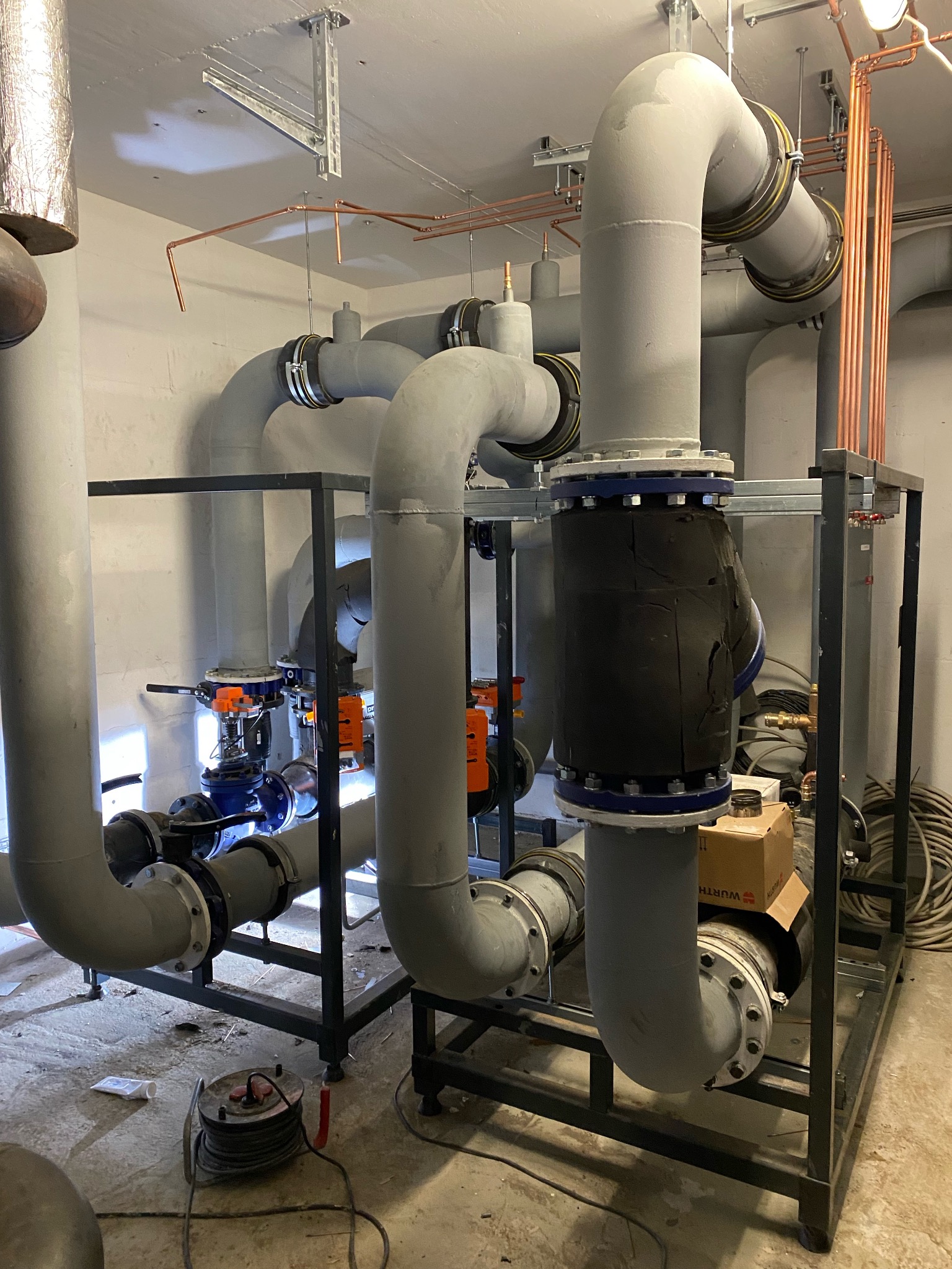 Złożona instalacja przemysłowa z szarych rur, zaworów i czarnej izolacji, wsparta na metalowej konstrukcji. Miedziane rurki pod sufitem. Widoczne narzędzia na podłodze.