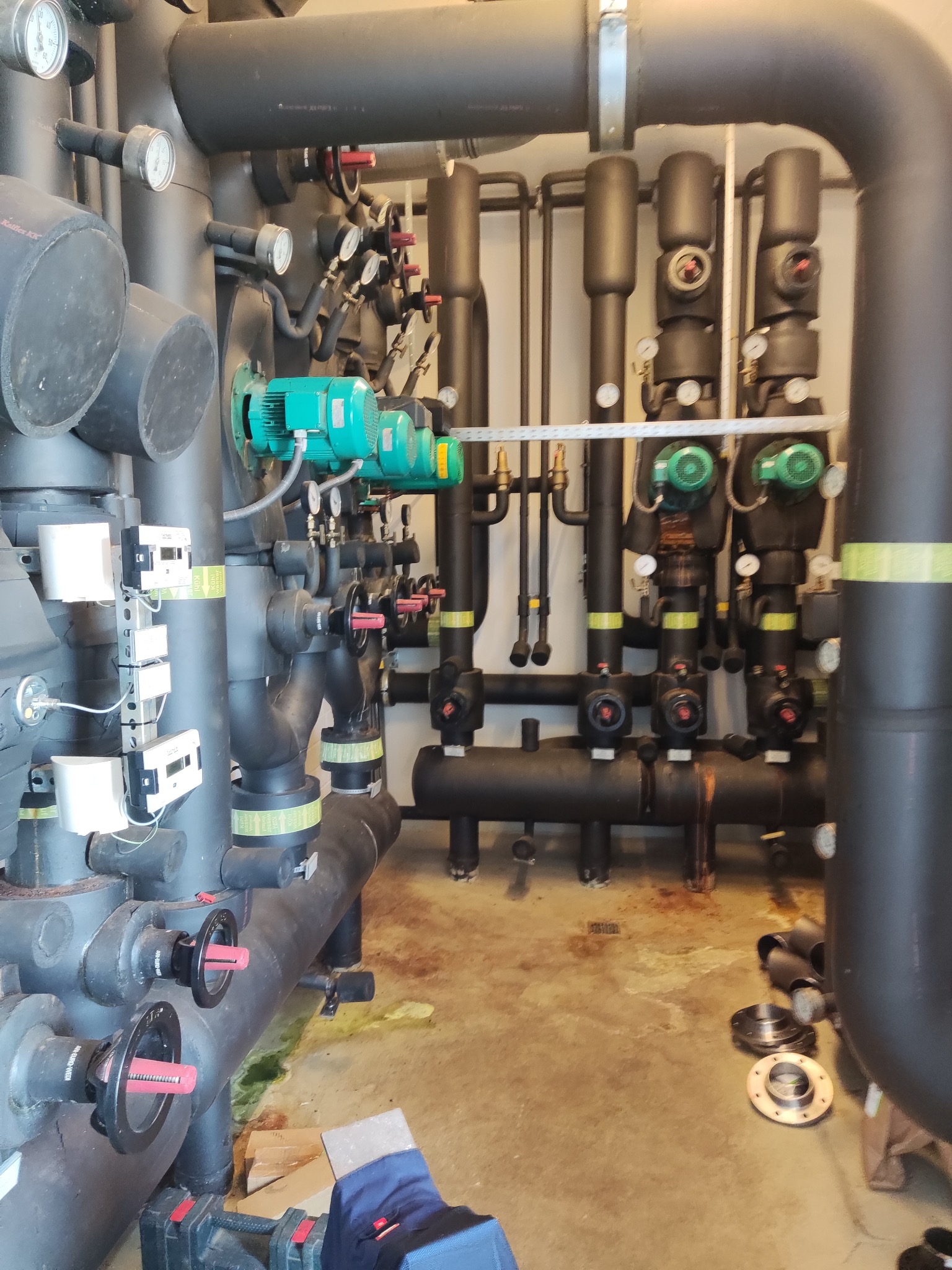 Złożona instalacja grzewcza z rurami, zaworami, manometrami i zielonymi pompami obiegowymi. Widoczne narzędzia i elementy montażowe na posadzce.