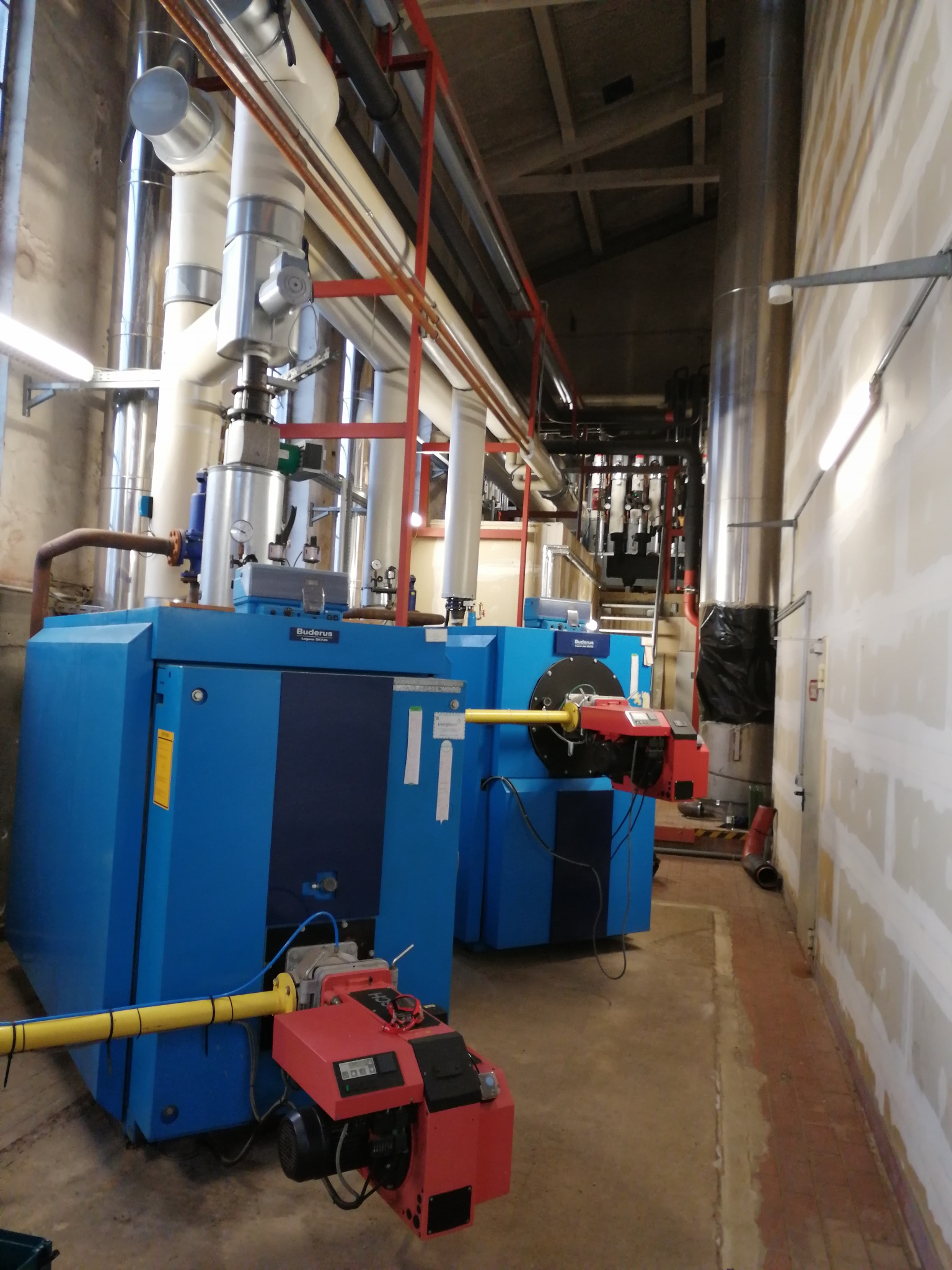 Dwa niebieskie, przemysłowe kotły Buderus z czerwonymi palnikami w kotłowni. Widoczne rury, instalacje i oświetlenie. Ściany częściowo wykończone, sufit z drewnianą konstrukcją.