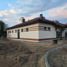 DEBKOBUD - Elewacja budynku jednokondygnacyjnego z brązowymi pasami i wykończeniem, widoczna z poziomu gruntu. Częściowo zagospodarowany teren wokół budynku, rusztowania przy ścianach.