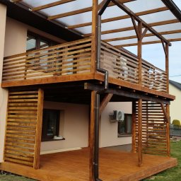 DEBKOBUD - Dwupoziomowy taras drewniany z zadaszeniem z poliwęglanu, brązowe drewno, widok z dołu, elementy konstrukcyjne, trawnik, fragment budynku w tle.