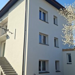 DEBKOBUD - Jasna, trzypiętrowa elewacja budynku z betonowymi schodami zewnętrznymi i kwitnącym drzewem. Widoczne okna z białymi ramami i ciemny dach.