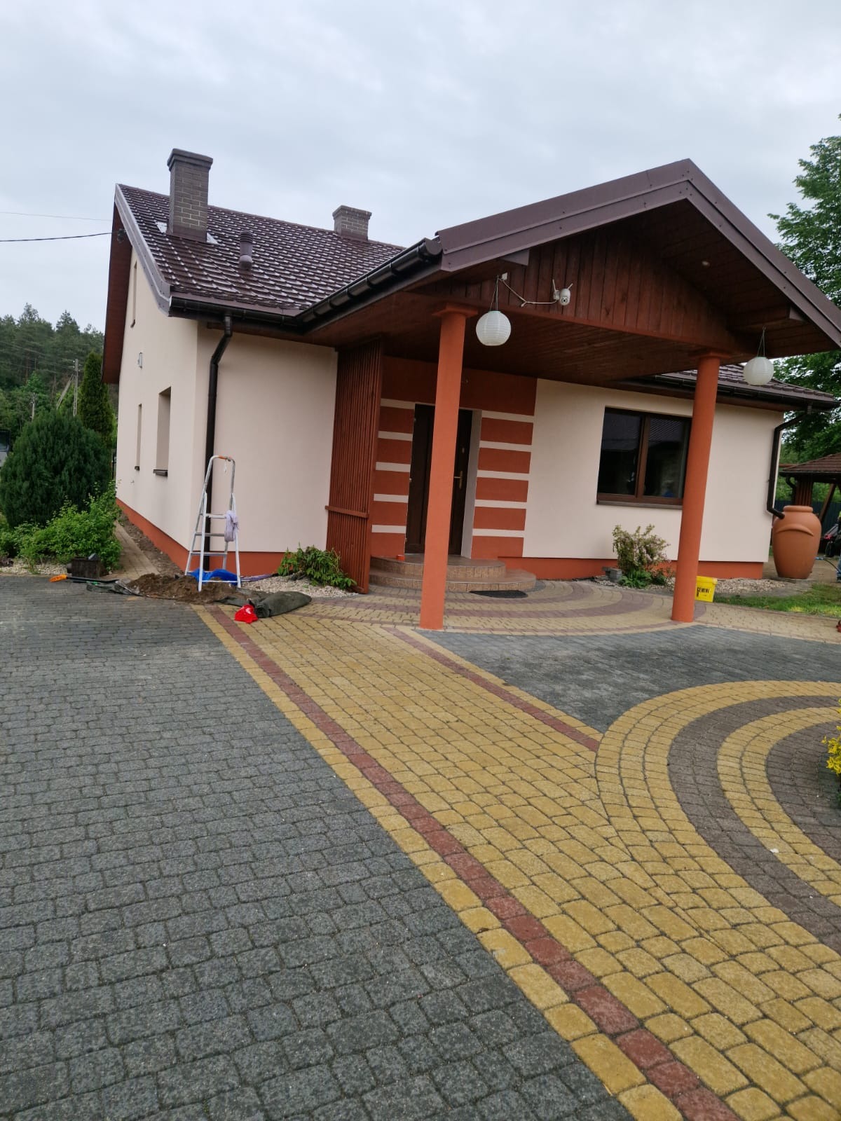 Jasny dom z brązowym dachem i podbitką, widoczne elementy elewacji, drabina oparta o ścianę, kostka brukowa na podjeździe. Prace wykończeniowe.