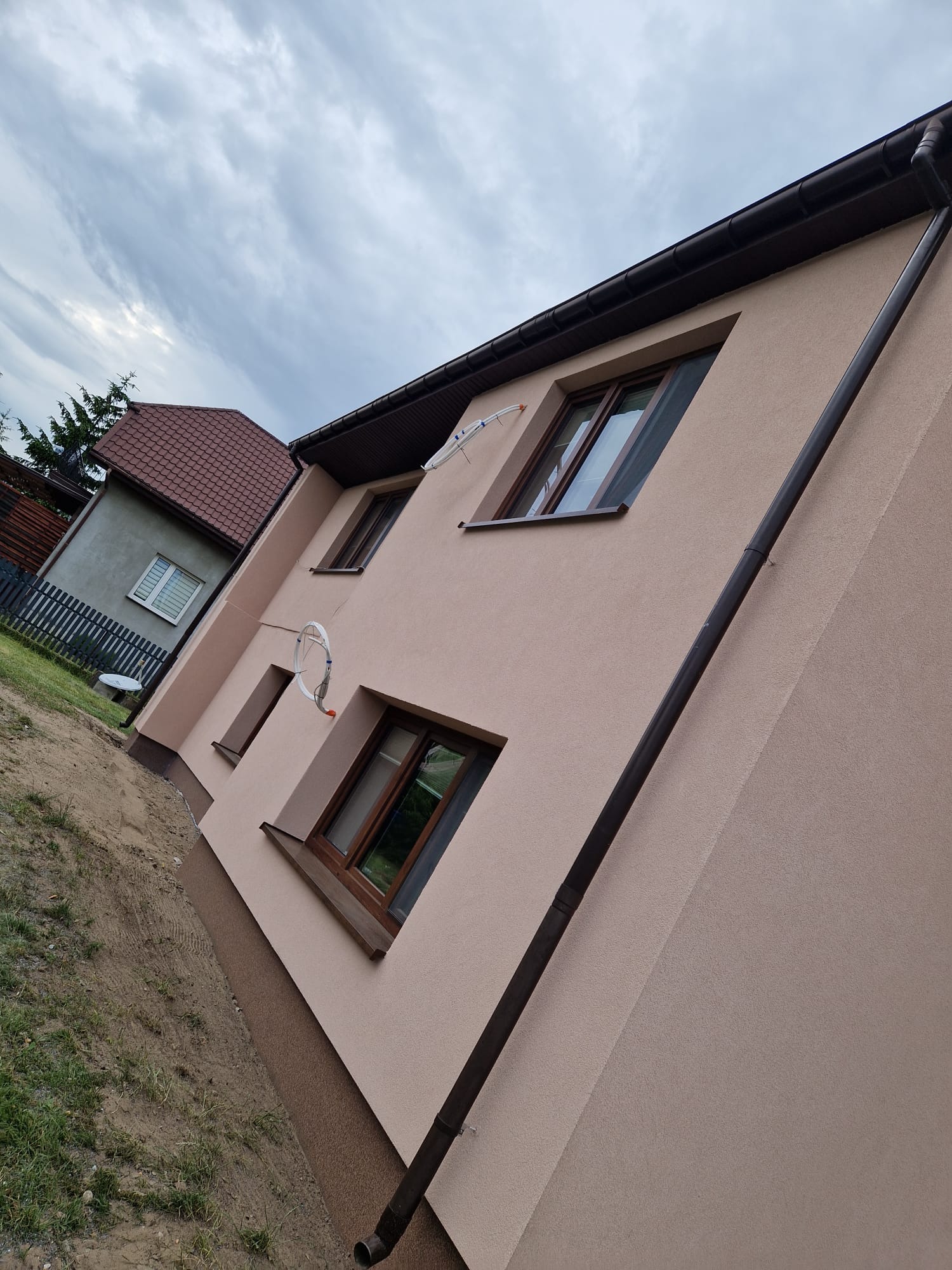 Elewacja domu jednorodzinnego w odcieniach beżu, z brązowymi oknami i rynnami. Widoczne detale wykończenia, antena satelitarna i fragment sąsiedniego budynku.