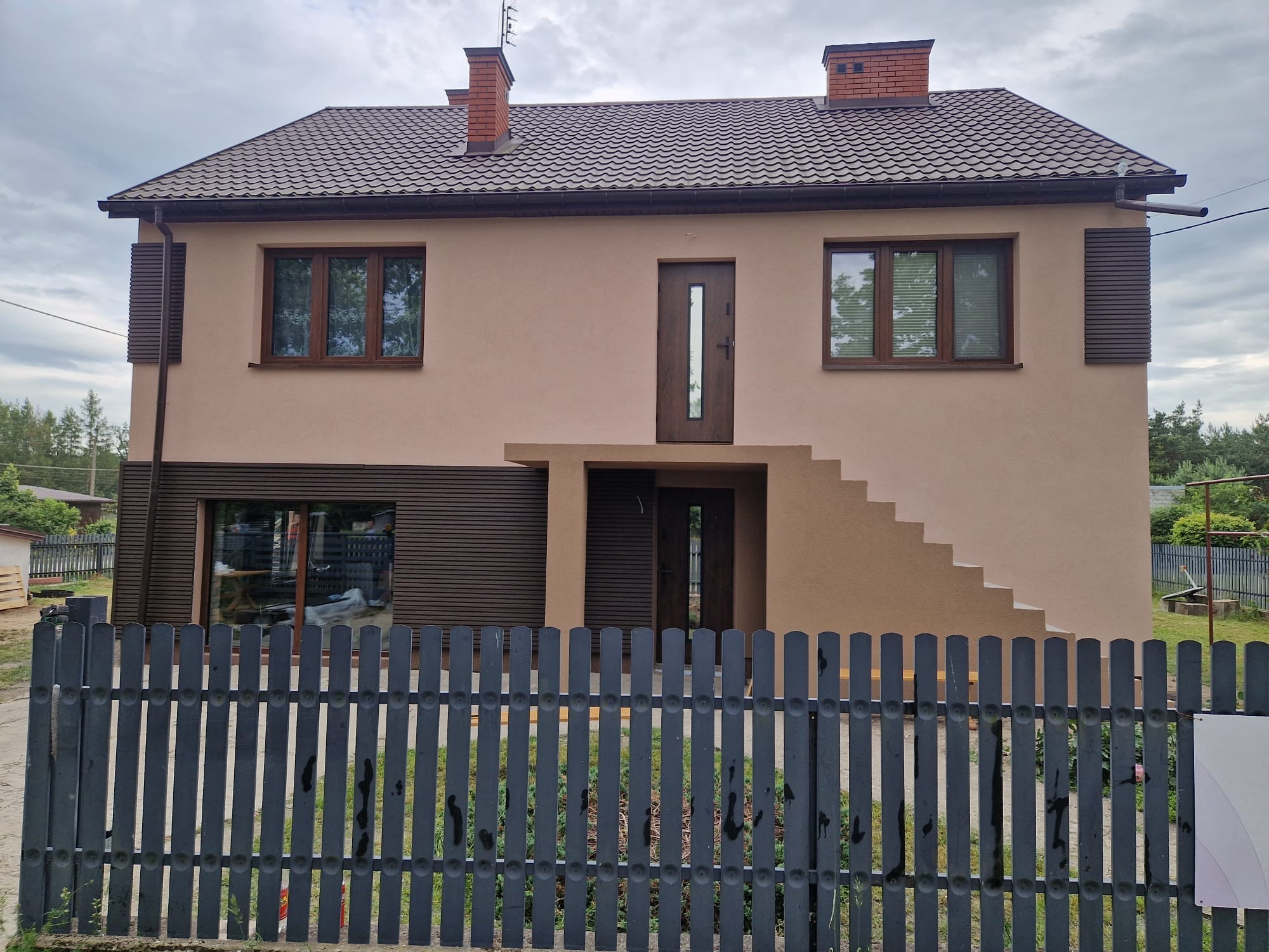 Dwukondygnacyjny dom z beżową elewacją, brązowymi oknami i dachem, widziany zza szarego płotu. Schody prowadzą do drzwi na piętrze.
