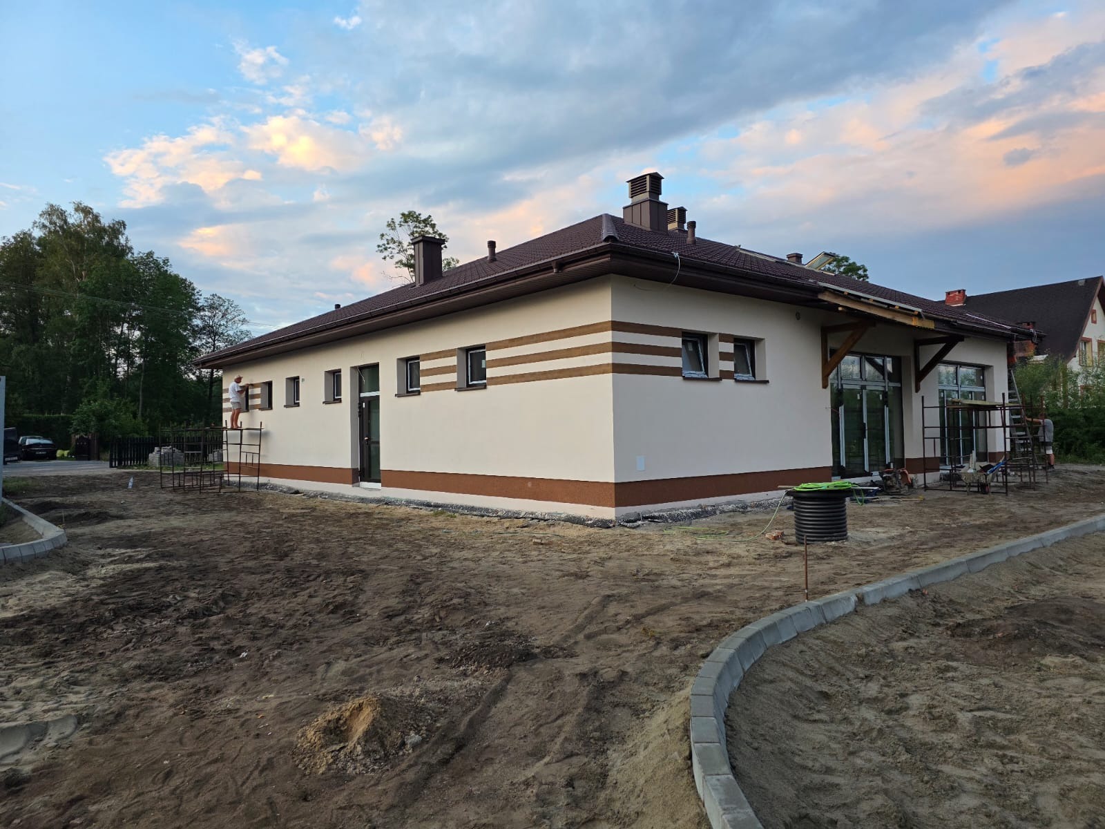 Elewacja budynku jednokondygnacyjnego z brązowymi pasami i wykończeniem, widoczna z poziomu gruntu. Częściowo zagospodarowany teren wokół budynku, rusztowania przy ścianach.