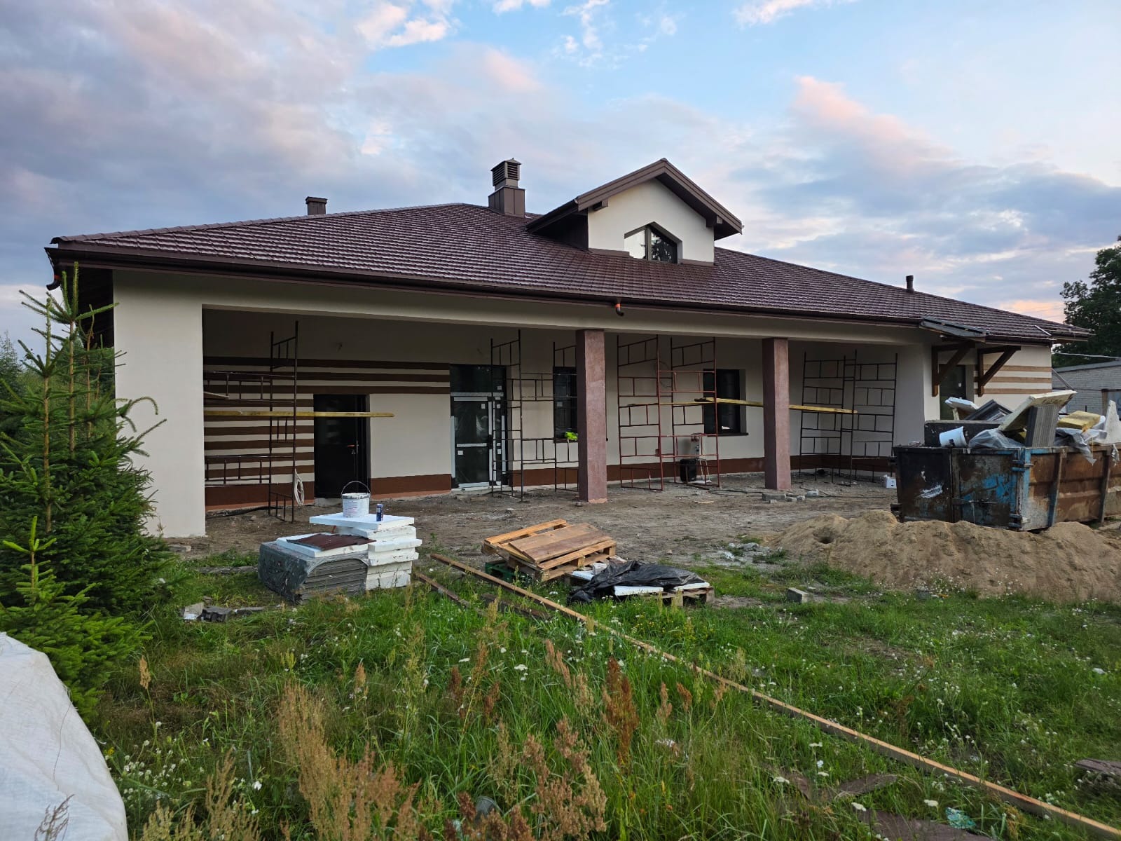 Dom jednorodzinny w trakcie prac elewacyjnych, widoczne rusztowania, materiały budowlane i częściowo otynkowana fasada. Brązowy dach i beżowe ściany.