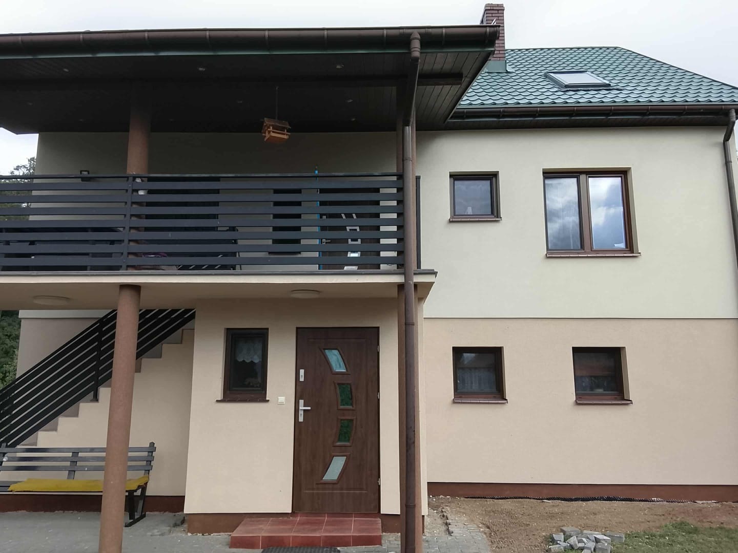 Dwukondygnacyjny dom z balkonem i zielonym dachem. Elewacja w dwóch odcieniach beżu, brązowe okna i drzwi. Widoczna część ogrodu i ławka.