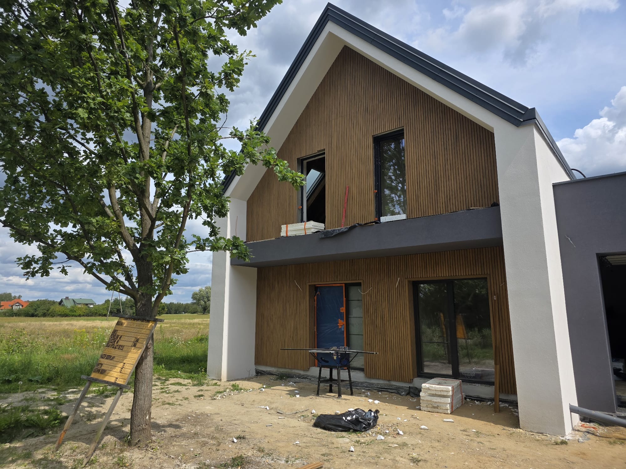 Nowoczesny dom jednorodzinny z drewnianą elewacją w trakcie montażu, widoczne narzędzia i materiały budowlane. Tablica informacyjna przy drzewie.
