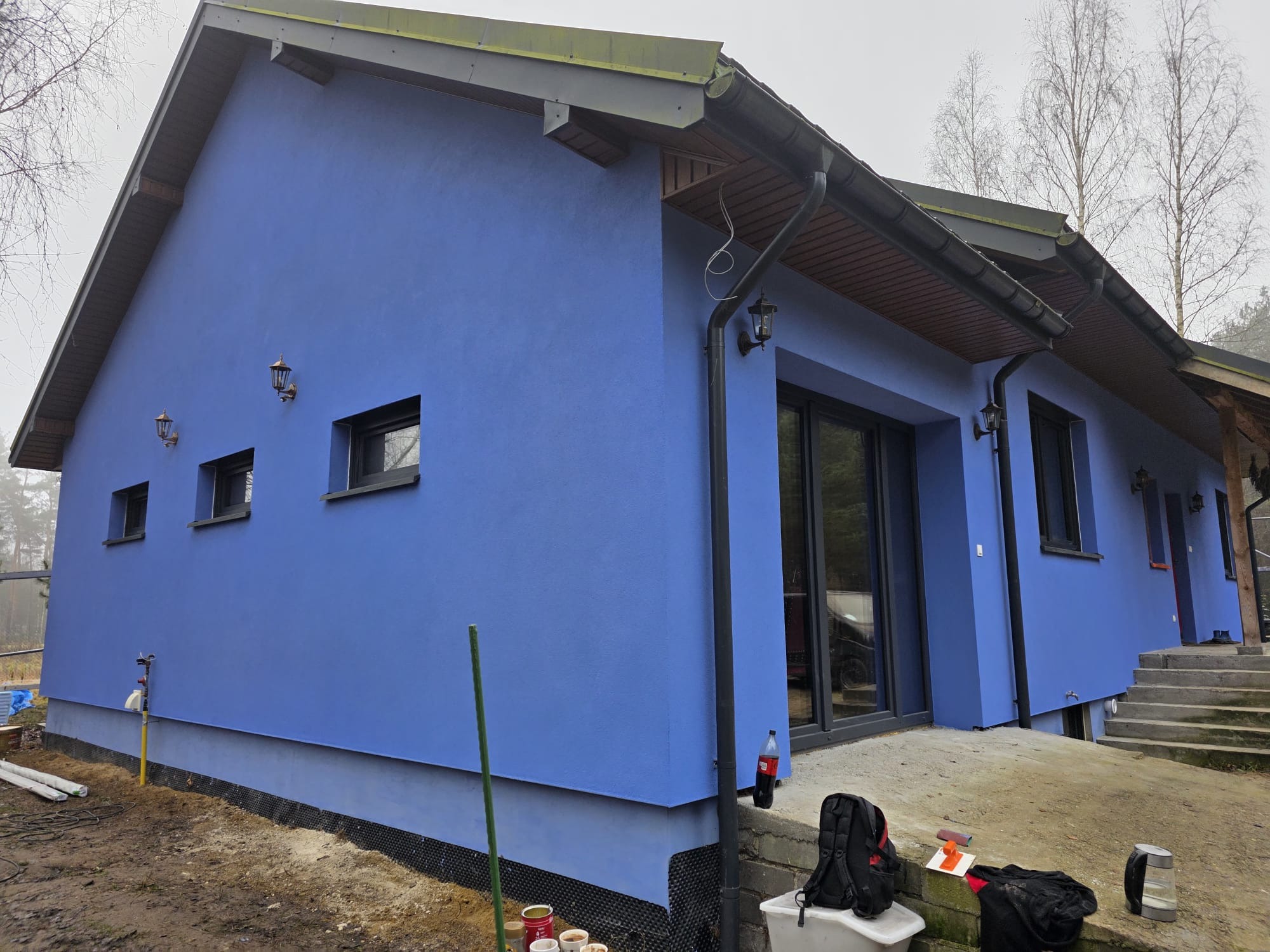 Niebieski dom jednorodzinny z ciemną stolarką okienną i oświetleniem zewnętrznym. Widoczny fragment elewacji, rynny i fragment dachu. Ujęcie z poziomu gruntu.