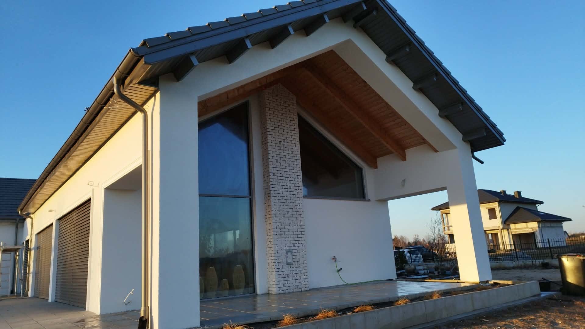 Nowoczesny dom jednorodzinny z białymi ścianami i ceglanym filarem, duże okna, widoczne elementy drewniane pod dachem, błękitne niebo w tle.