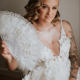 Przedsiębiorstwo Handlowo-Usługowe DAWID KREPS - Panna młoda w białej sukni z kwiatowymi aplikacjami, trzymająca wachlarz z napisem 'BRIDE', z widocznym tatuażem na ramieniu i delikatnym makijażem.