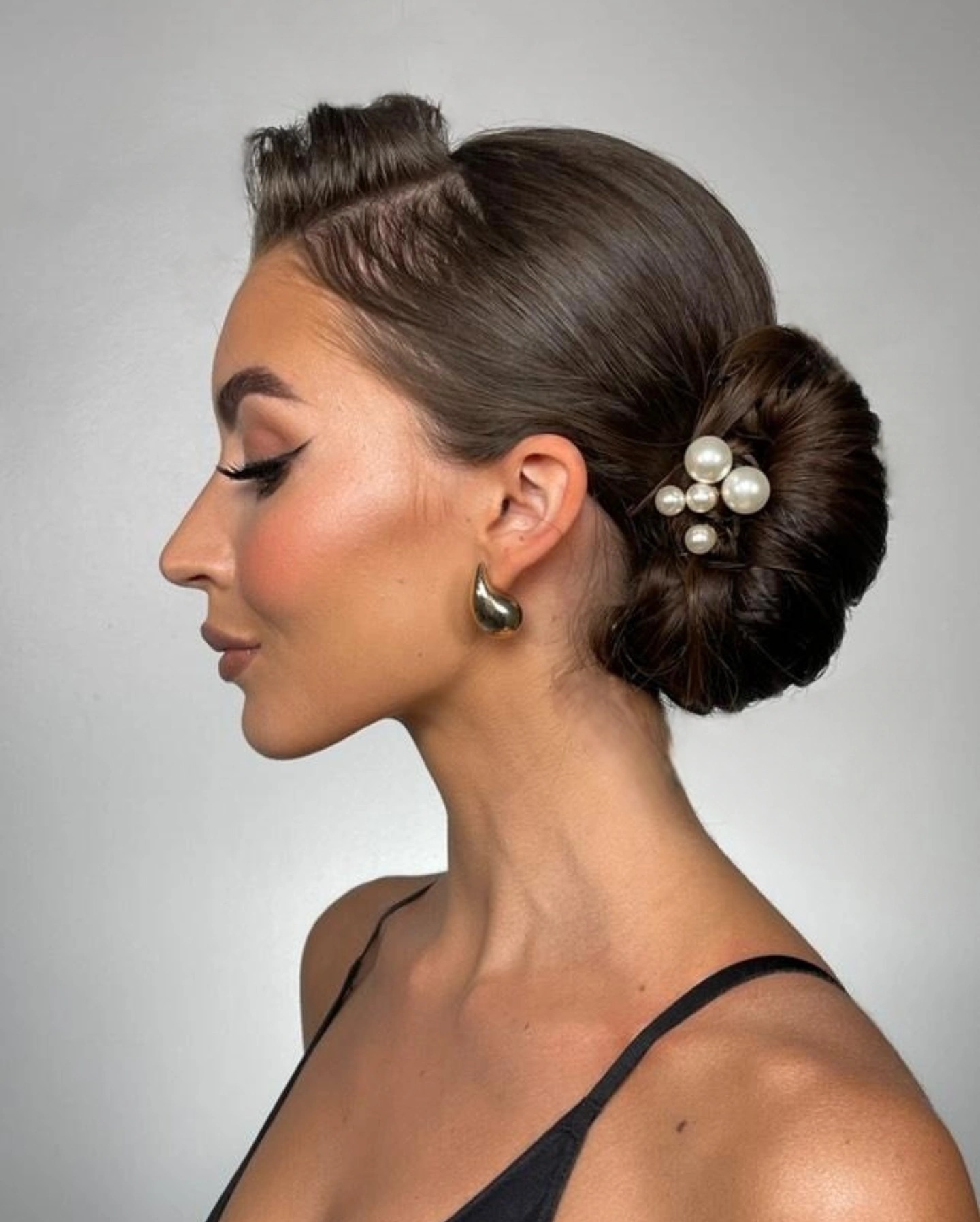 Elegancka fryzura upięta w kok z perłami, widok z profilu, podkreślone oko eyelinerem, złote kolczyki, gładko zaczesane włosy, czarny top na ramiączkach.