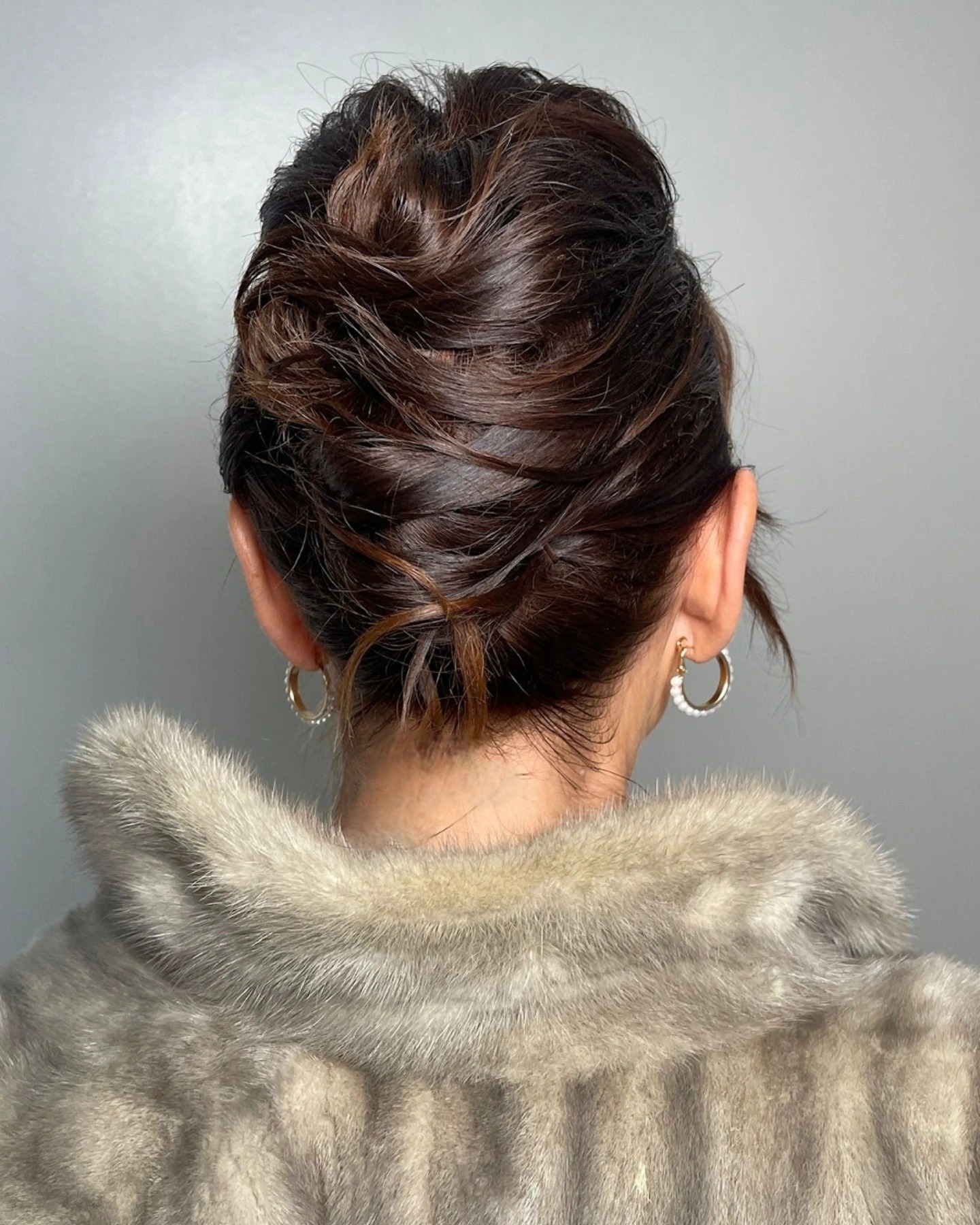 Elegancka fryzura upięta z tyłu głowy, widoczne złote kolczyki i futro w odcieniach szarości na tle szarej ściany. Upięcie włosów z bliska.