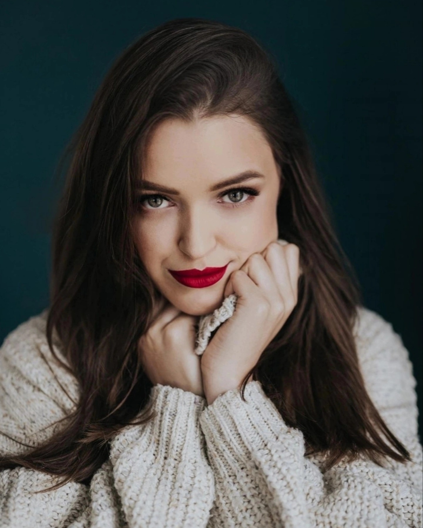 Portret kobiety z długimi brązowymi włosami, w kremowym swetrze, z czerwonymi ustami na ciemnym tle. Dłonie pod brodą, spojrzenie w obiektyw.