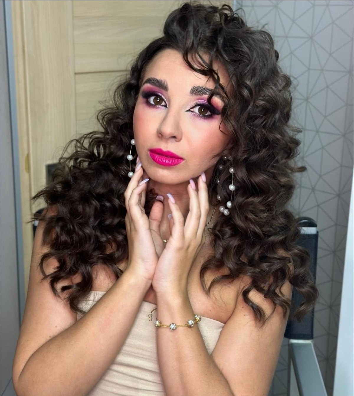 Portret kobiety z intensywnym makijażem oczu w odcieniach różu i fuksji, z burzą ciemnych loków i delikatną biżuterią, pozującej na krześle.