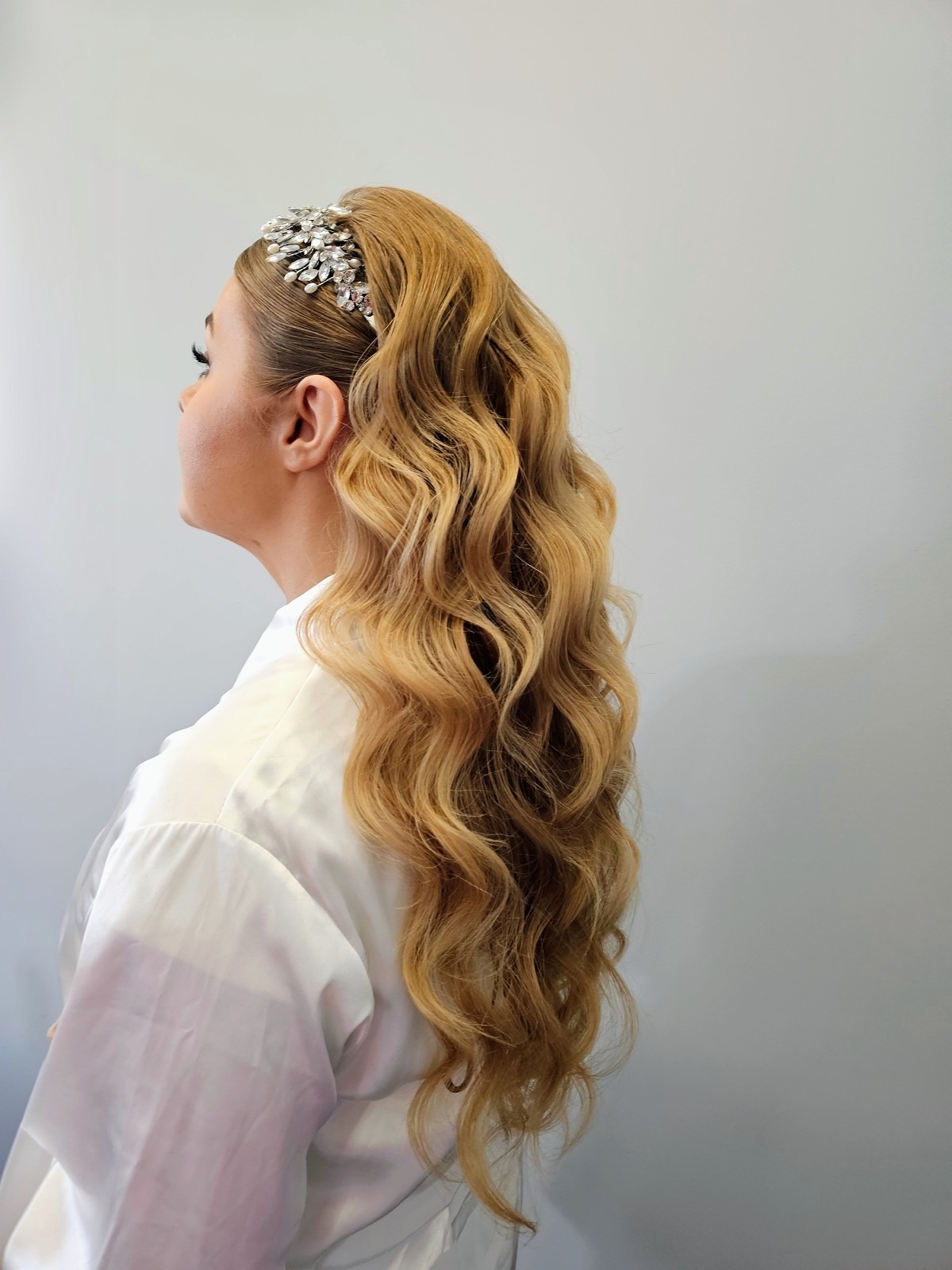 Profil kobiety z długimi, blond włosami ułożonymi w fale, ozdobionymi błyszczącą opaską. Ubrana w białą koszulę, na jasnym tle.