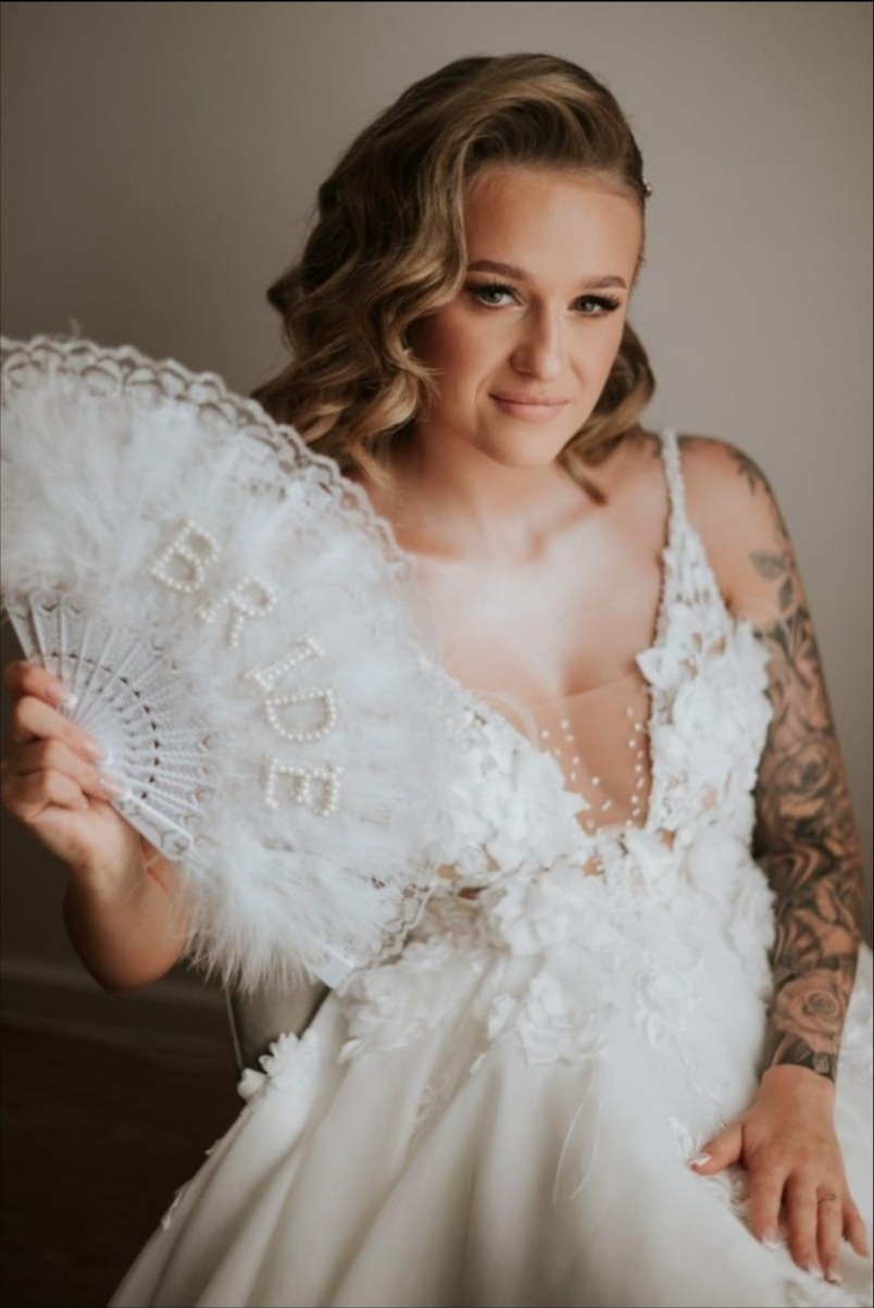 Panna młoda w białej sukni z kwiatowymi aplikacjami, trzymająca wachlarz z napisem 'BRIDE', z widocznym tatuażem na ramieniu i delikatnym makijażem.