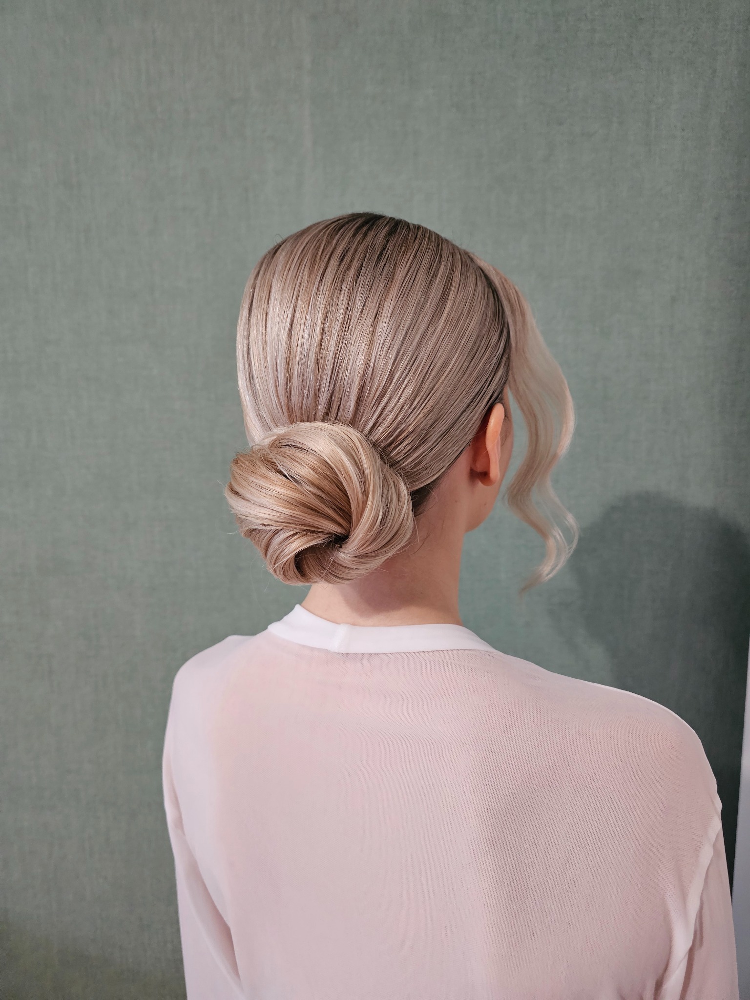 Elegancka fryzura upięta w kok z boku głowy, z delikatnym, opadającym pasmem. Jasne włosy na tle stonowanej, zielonej ściany. Widok z tyłu.
