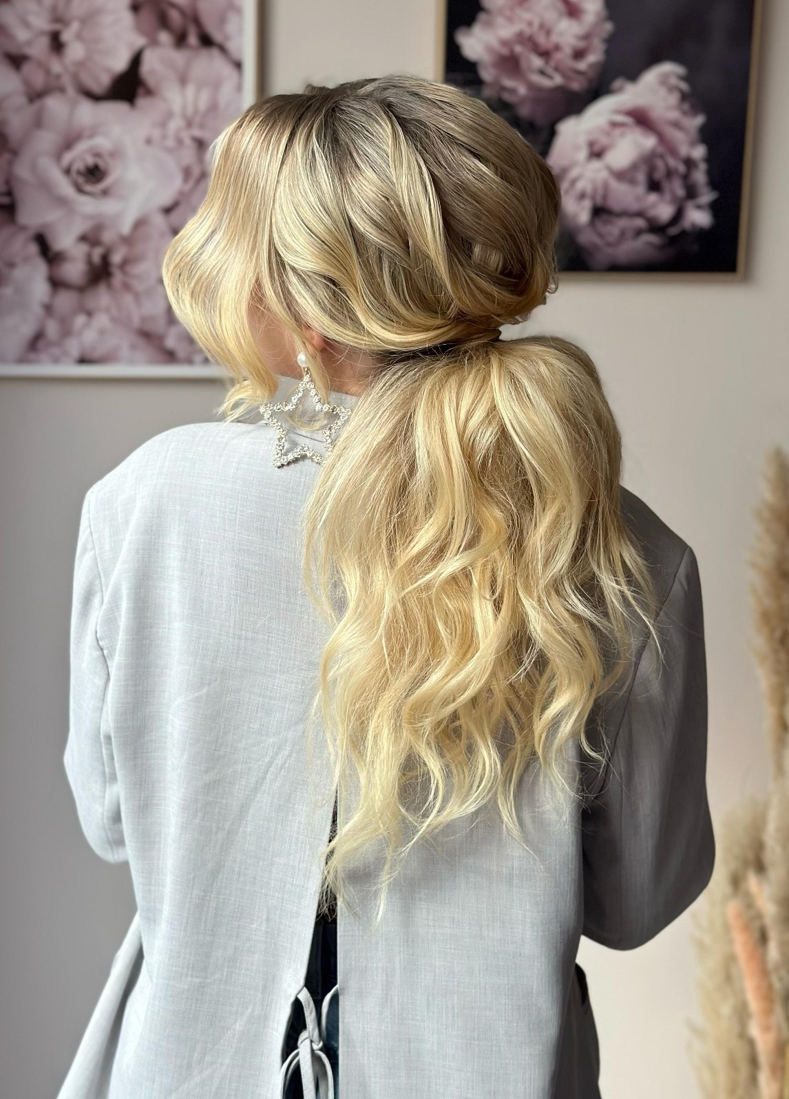 Blond włosy upięte w kok i kucyk, widok z tyłu, ozdobne kolczyki w kształcie gwiazd, szara marynarka, w tle kwiatowe obrazy i trawa pampasowa.