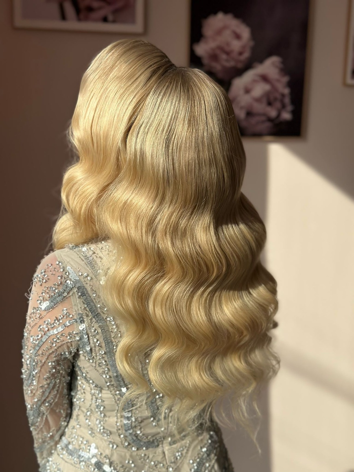 Elegancka fryzura blond z falami, widok z tyłu, na tle ściany z obrazami. Sukienka zdobiona cekinami. Delikatne światło podkreśla blask włosów.