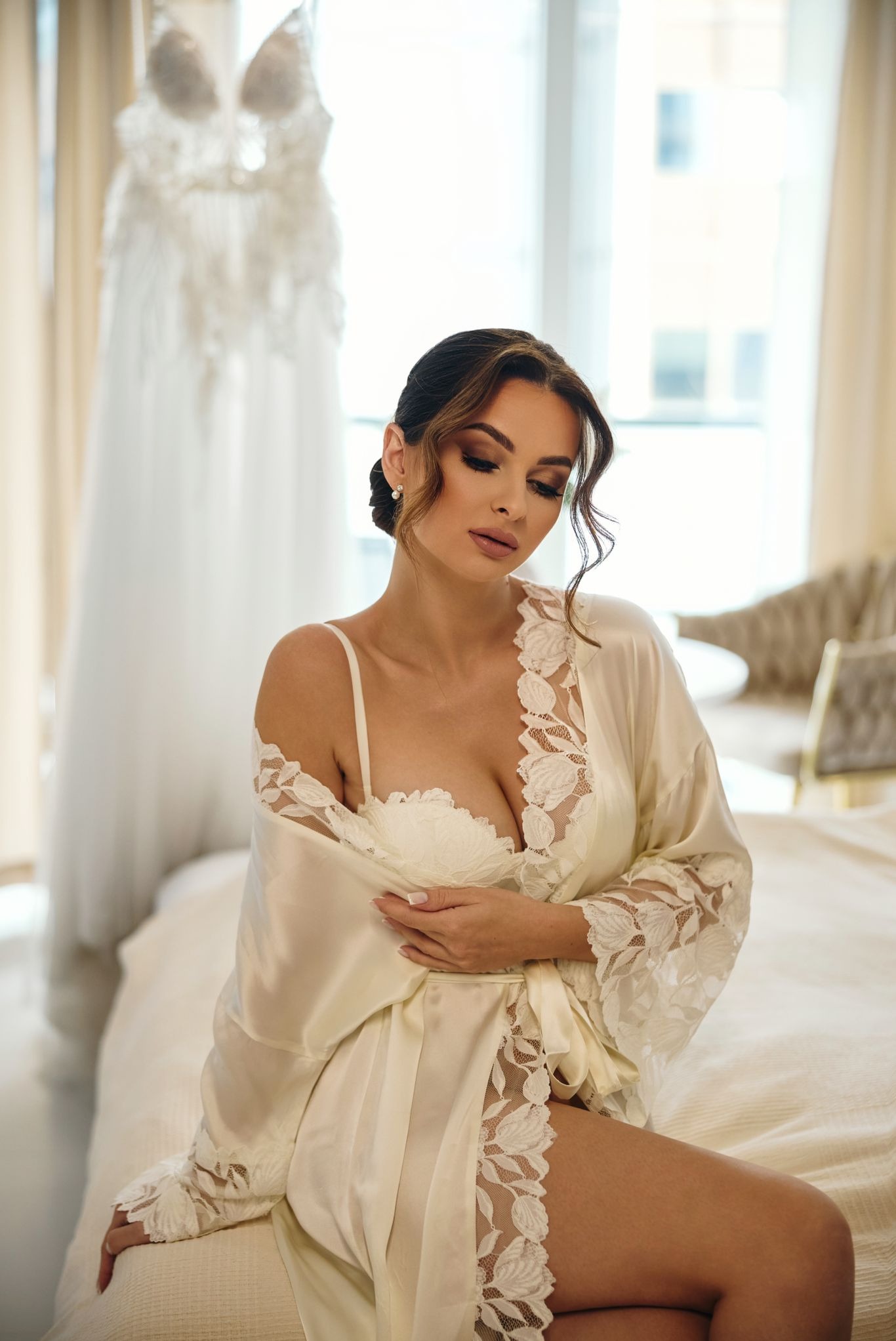 Kobieta w satynowym szlafroku z koronką, siedząca na łóżku. W tle suknia ślubna i jasne wnętrze. Subtelny makijaż i upięte włosy. Delikatne światło podkreśla urodę.