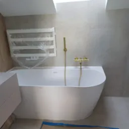 Nowoczesna łazienka w szarościach z białą wanną i złotą armaturą. Grzejnik drabinkowy owinięty folią ochronną. Umywalka nablatowa z szafką o ryflowanych frontach.