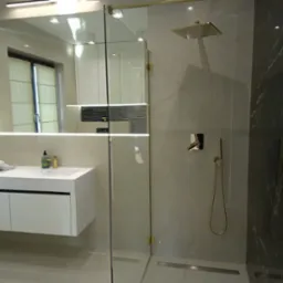Nowoczesna łazienka z kabiną prysznicową typu walk-in, armaturą w kolorze złota i białą szafką pod umywalką. Widoczne marmurowe płytki i minimalistyczne oświetlenie.