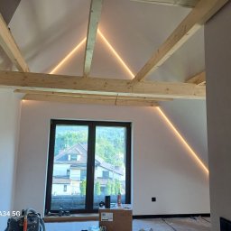 FIRMA REMONTOWA FINISZ SIKORA WIESŁAW - Poddasze po remoncie: odsłonięte drewniane belki stropowe z dekoracyjnym oświetleniem LED, świeżo pomalowane ściany, widok na zewnątrz przez duże okno.