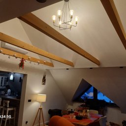 FIRMA REMONTOWA FINISZ SIKORA WIESŁAW - Jasne wnętrze poddasza z drewnianymi belkami stropowymi i nowoczesnym żyrandolem. Widoczny fragment kuchni w tle, oświetlony lampą stojącą.