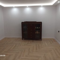 FIRMA REMONTOWA FINISZ SIKORA WIESŁAW - Pokój po remoncie: parkiet ułożony w jodełkę, białe ściany z listwami przypodłogowymi, oświetlenie sufitowe LED i ciemny kredens z przeszklonymi drzwiczkami.