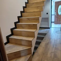 Drewniane schody z czarnymi krawędziami, prowadzące w górę. Podłoga wyłożona panelami w podobnym odcieniu drewna. Widoczne drzwi wejściowe.