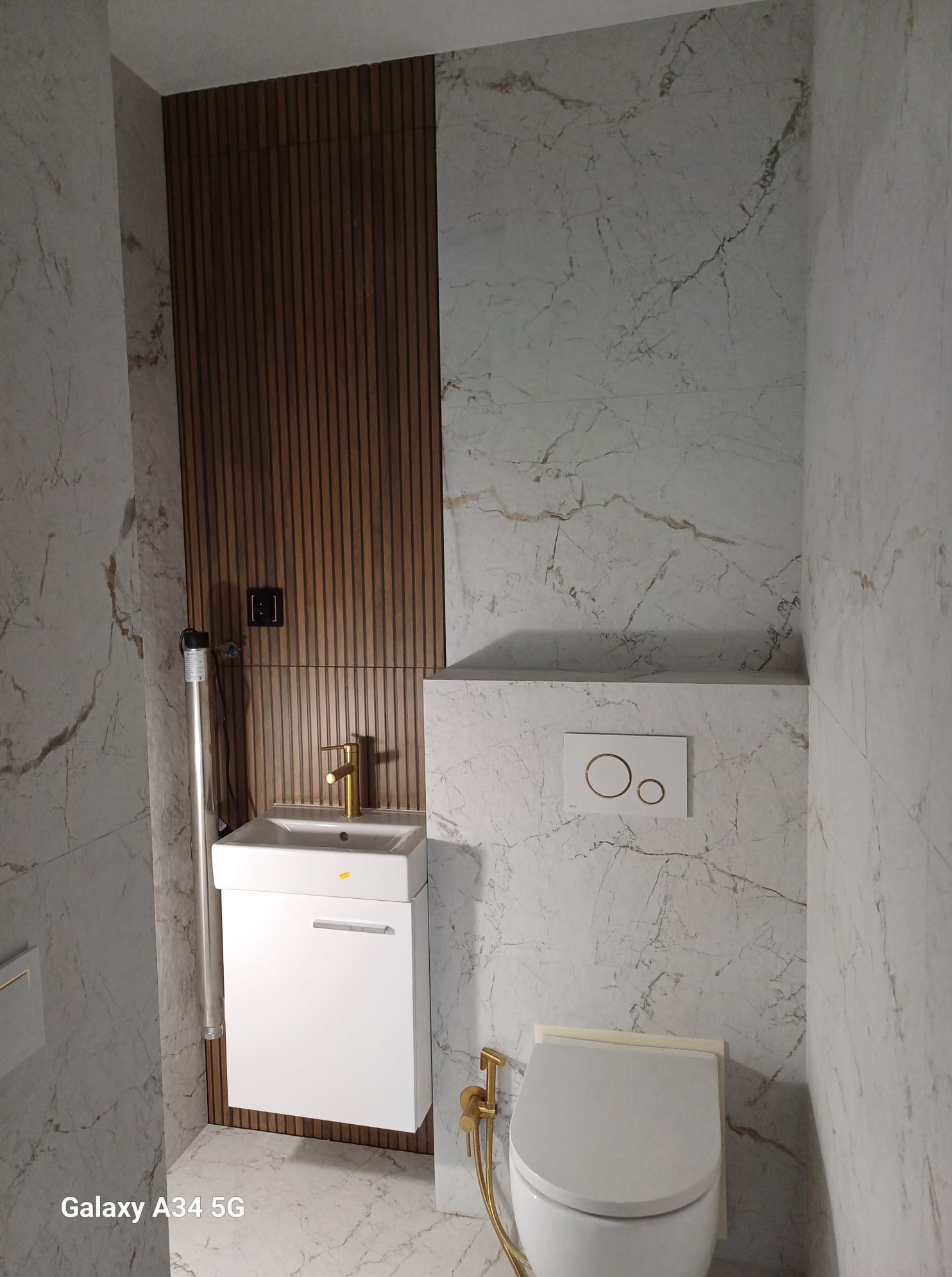Nowoczesna łazienka z marmurowymi płytkami, drewnianą ścianą, minimalistyczną umywalką z szafką i toaletą z złotymi akcentami. Wykończenie z dbałością o detale.