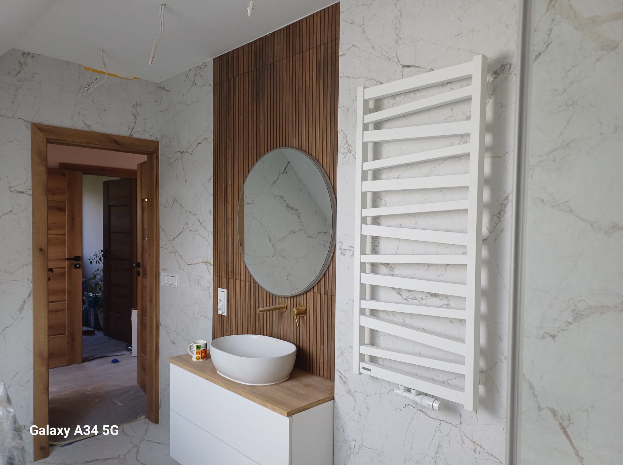 Nowoczesna łazienka z umywalką nablatową, lustrem w okrągłej ramie, grzejnikiem drabinkowym i drewnianymi panelami na ścianie z marmurowym wzorem. Widoczne niedokończone wykończenie.