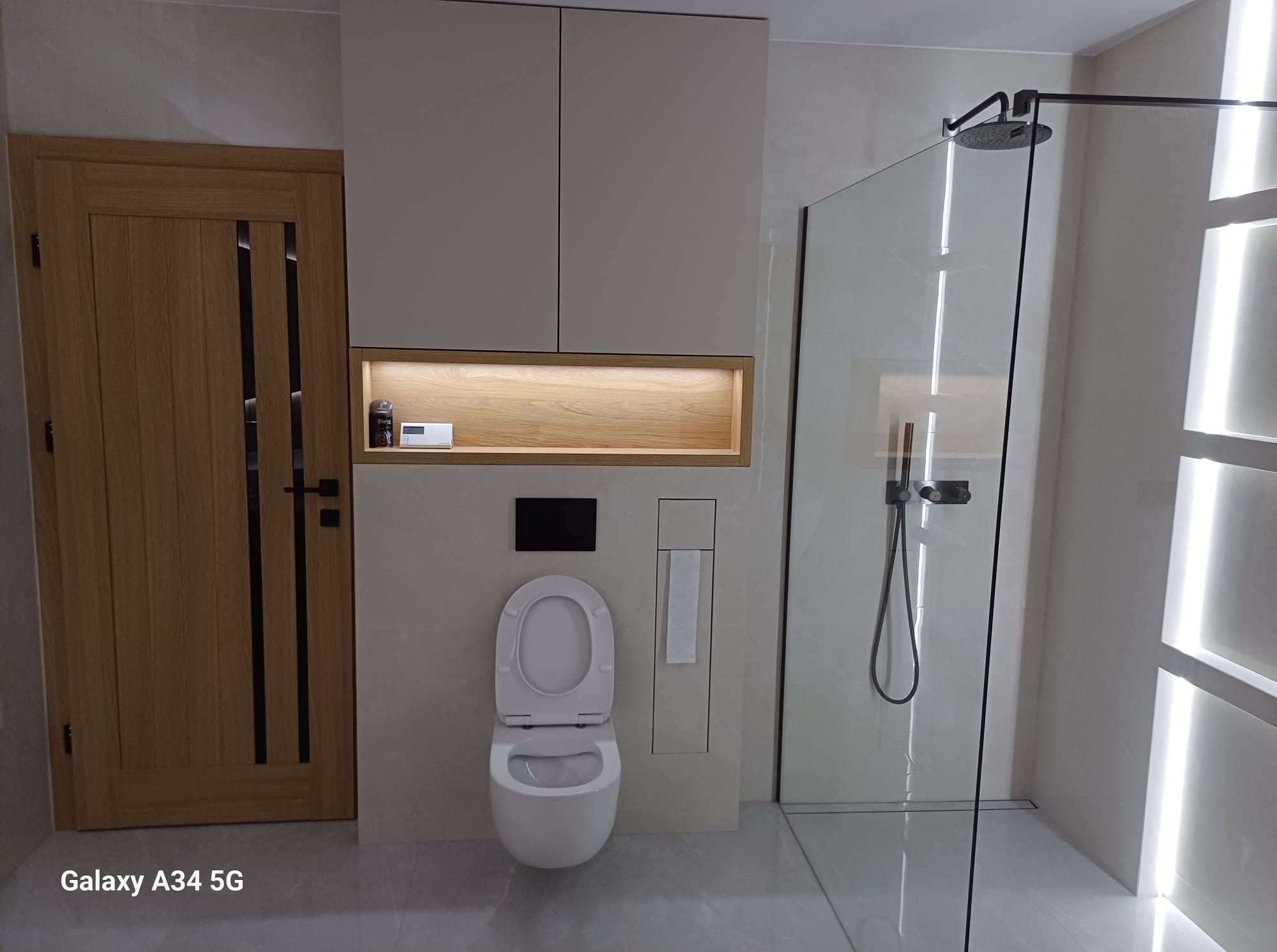 Nowoczesna łazienka z kabiną prysznicową typu walk-in, podwieszaną toaletą i szafkami w jasnych odcieniach. Drewniane akcenty i minimalistyczne oświetlenie tworzą spójną całość.