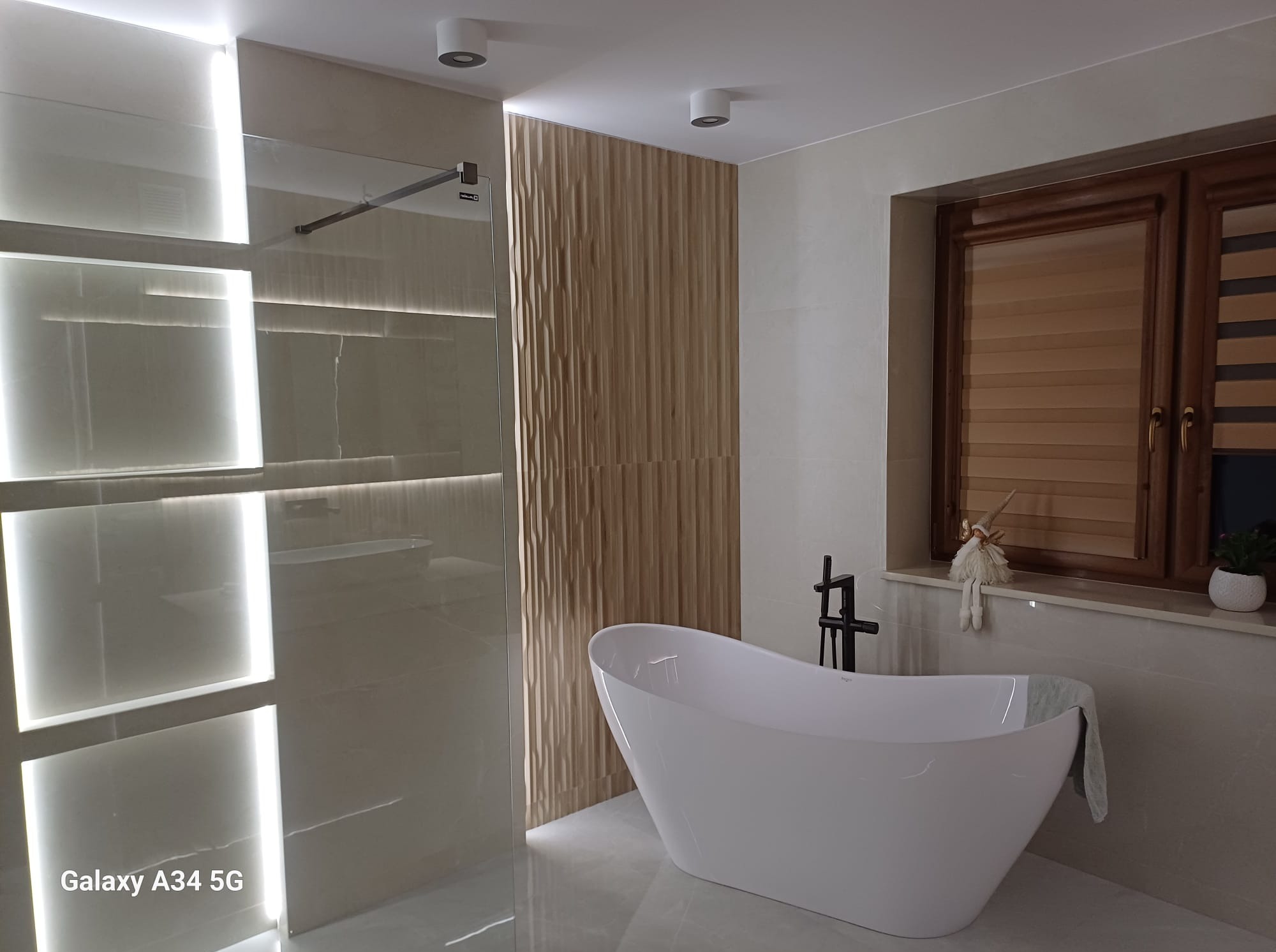 Nowoczesna łazienka z wolnostojącą wanną, podświetlaną kabiną prysznicową i dekoracyjną ścianą z paneli. Okno z drewnianą ramą i roletami.