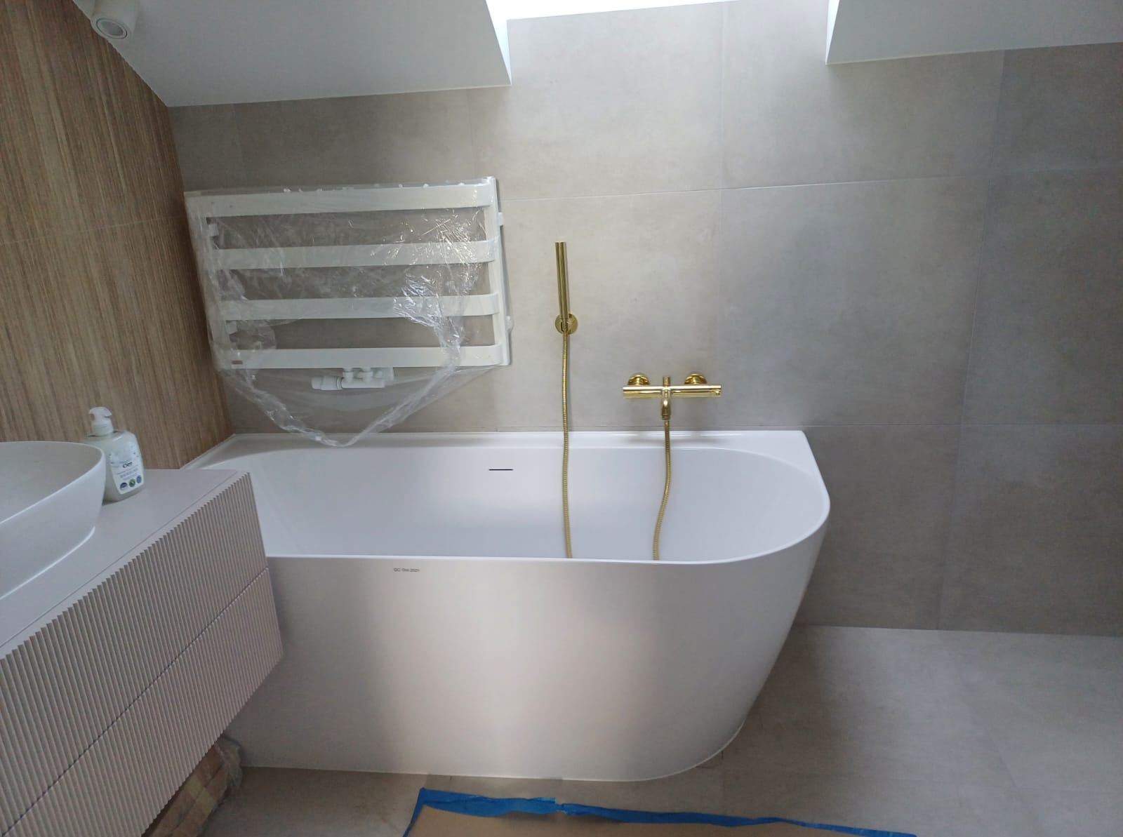 Nowoczesna łazienka w szarościach z białą wanną i złotą armaturą. Grzejnik drabinkowy owinięty folią ochronną. Umywalka nablatowa z szafką o ryflowanych frontach.