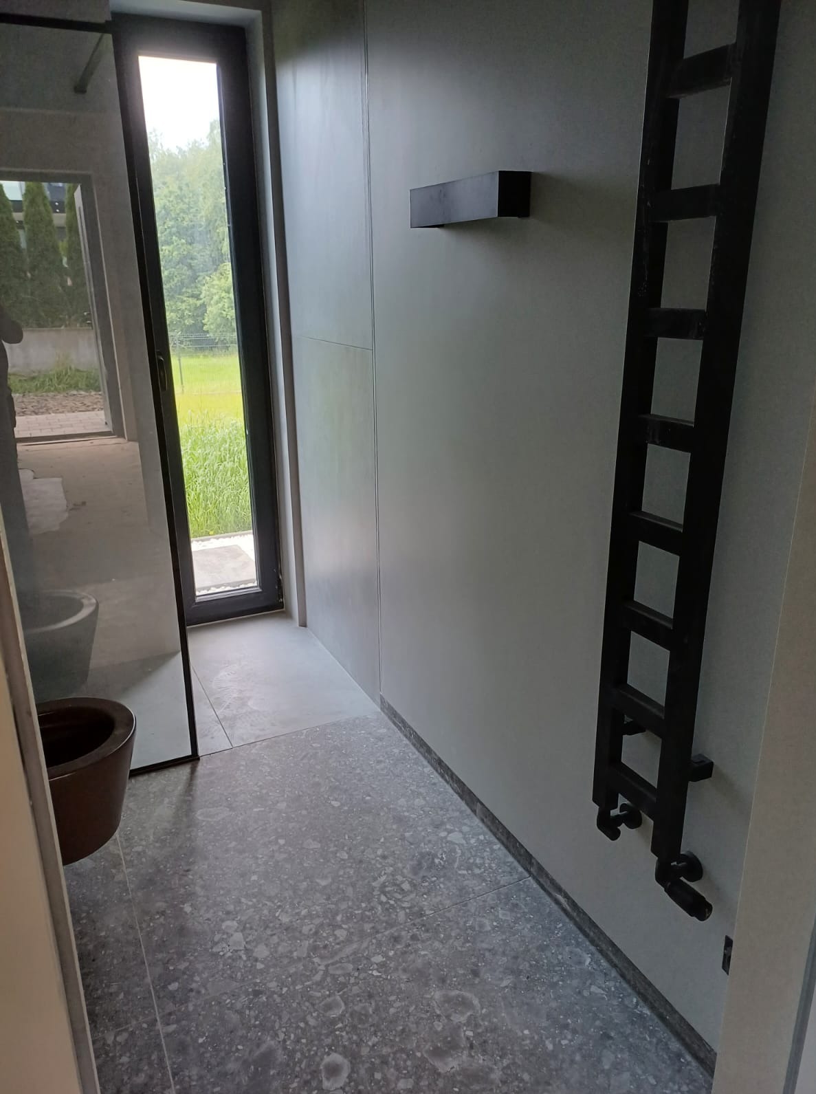 Minimalistyczna łazienka z szarą terazzo posadzką, czarnym grzejnikiem drabinkowym i oknem z widokiem na zieleń. Uwagę przykuwa brązowa miska toaletowa.