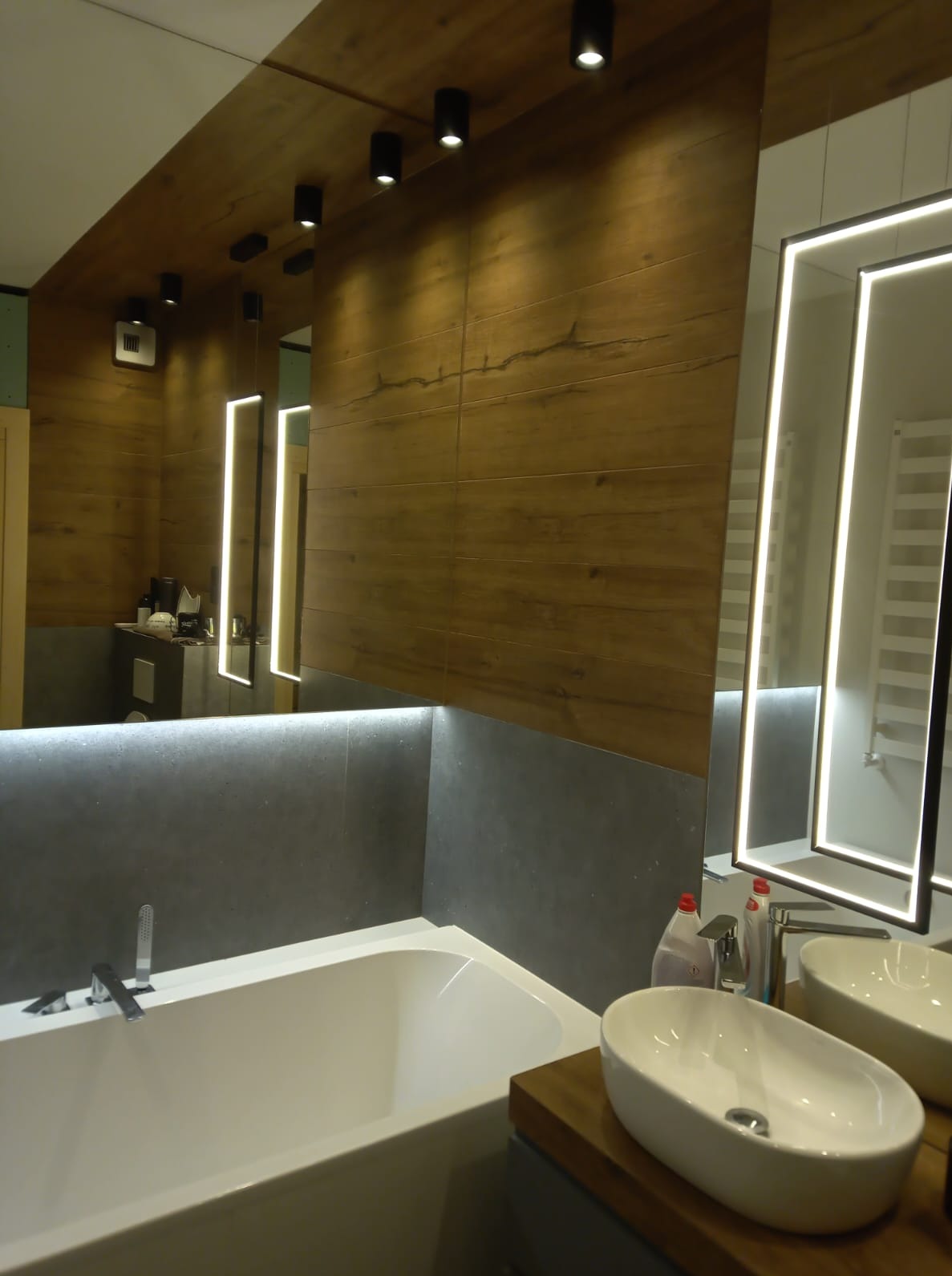 Nowoczesna łazienka z drewnopodobnymi płytkami, podświetlanym lustrem i szarą zabudową wanny. Minimalistyczny design z armaturą w chromie.