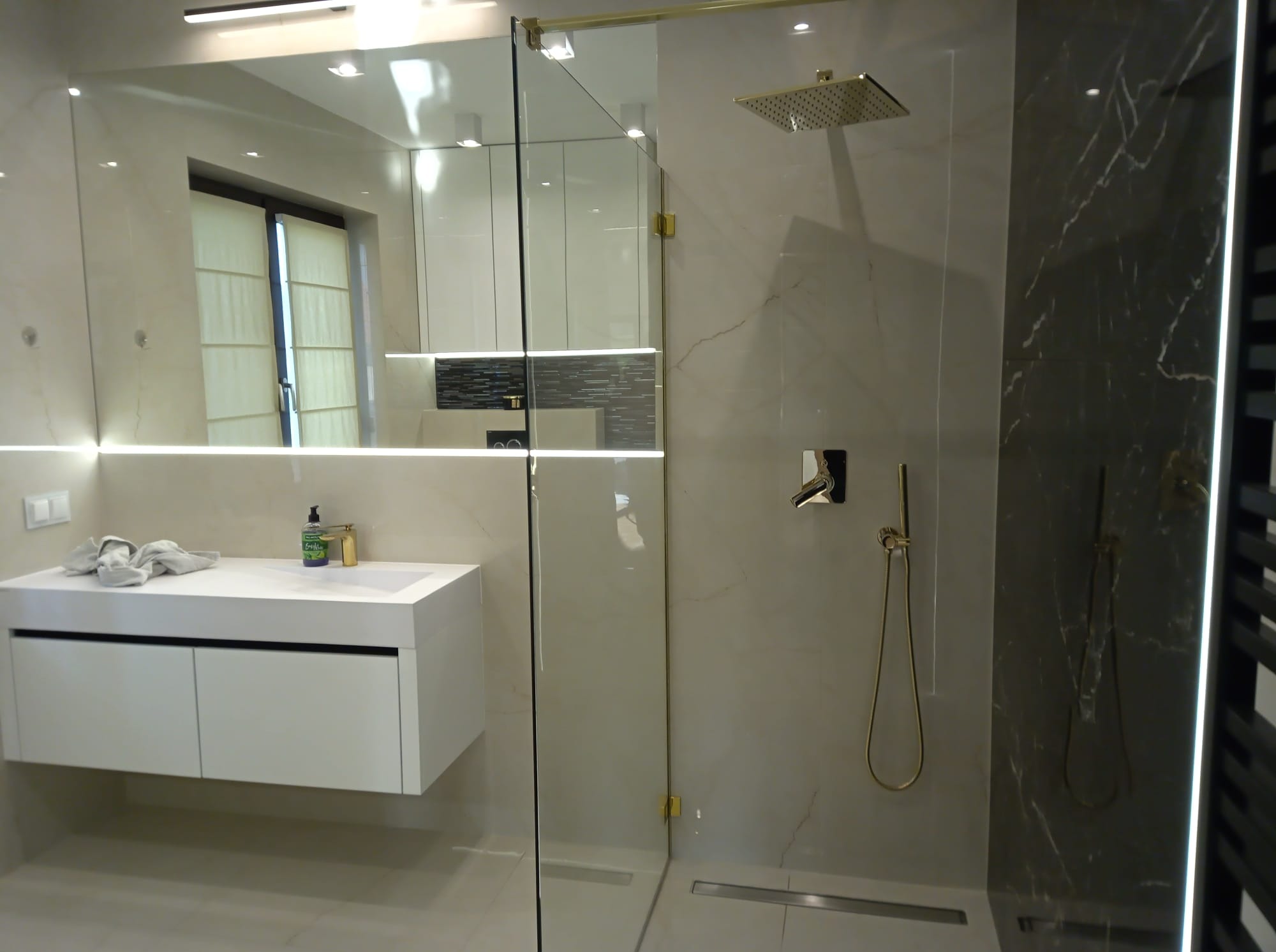 Nowoczesna łazienka z kabiną prysznicową typu walk-in, armaturą w kolorze złota i białą szafką pod umywalką. Widoczne marmurowe płytki i minimalistyczne oświetlenie.