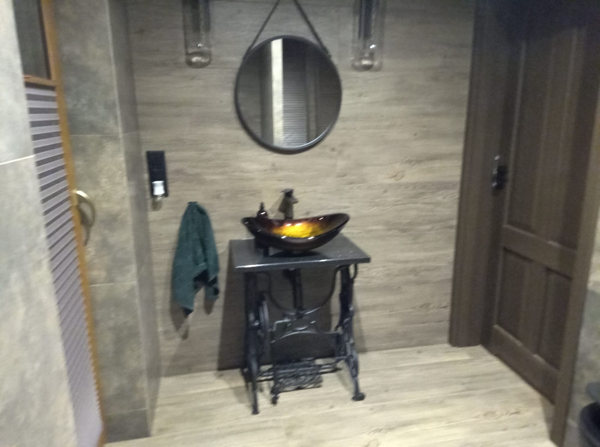 Umywalka w stylu retro z misą w odcieniach brązu i żółci, umieszczona na metalowym stelażu przypominającym maszynę do szycia. Lustro w okrągłej ramie.