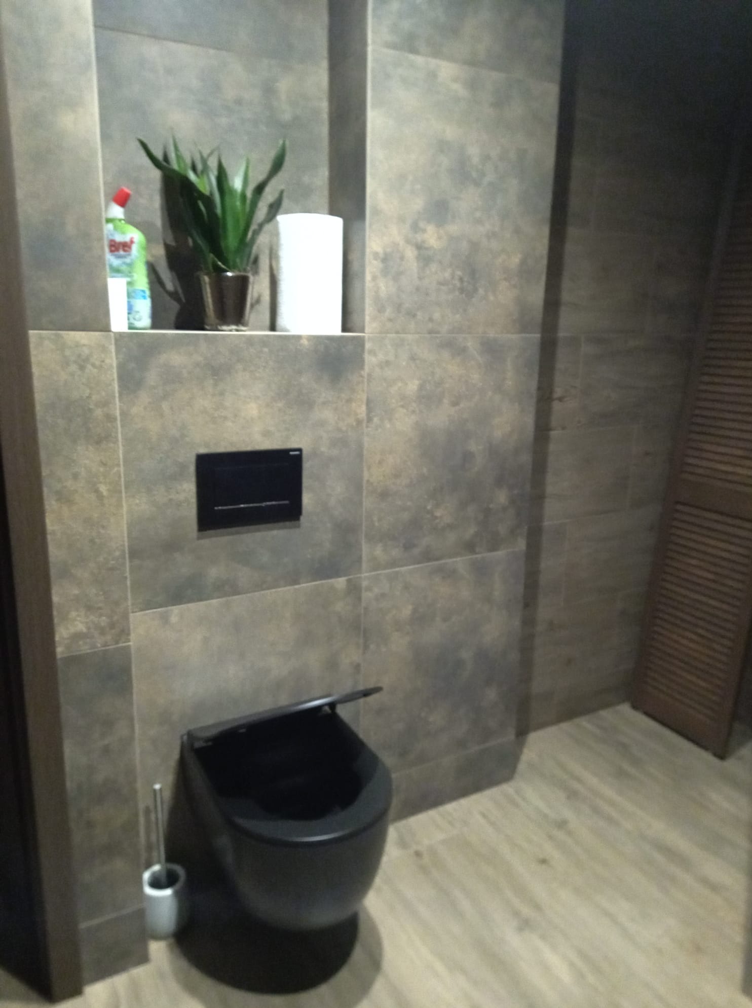 Nowoczesna toaleta w łazience z szarymi płytkami, czarną miską wiszącą i roślinami w niszy. Widoczny fragment drzwi z żaluzjami. Minimalistyczny design.