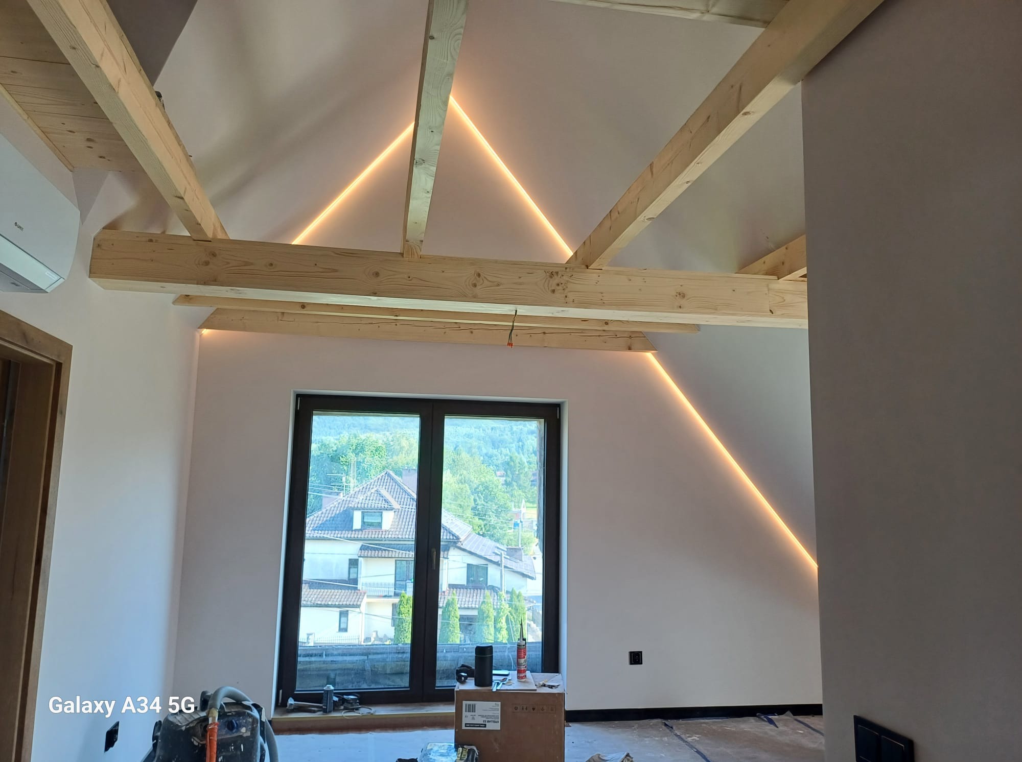 Poddasze po remoncie: odsłonięte drewniane belki stropowe z dekoracyjnym oświetleniem LED, świeżo pomalowane ściany, widok na zewnątrz przez duże okno.